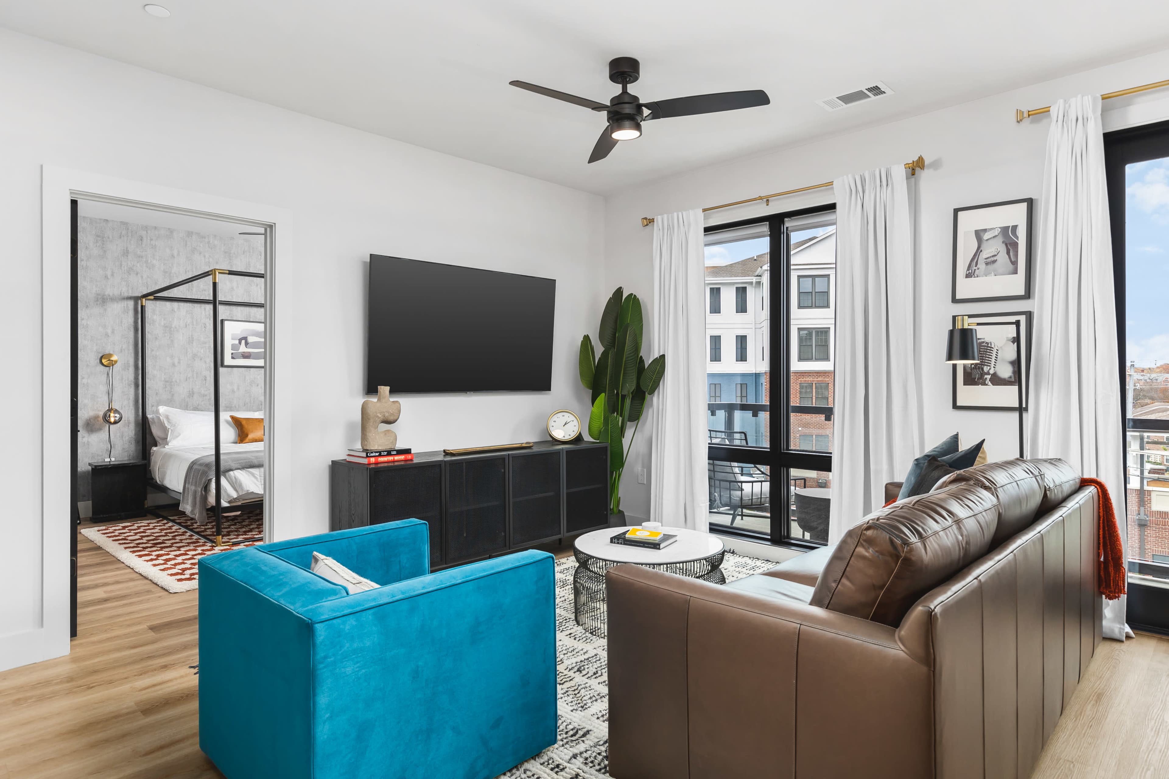 A modern living room features a brown couch, a turquoise armchair, a black TV stand, and large windows with white curtains, leading to a bedroom area in the background.