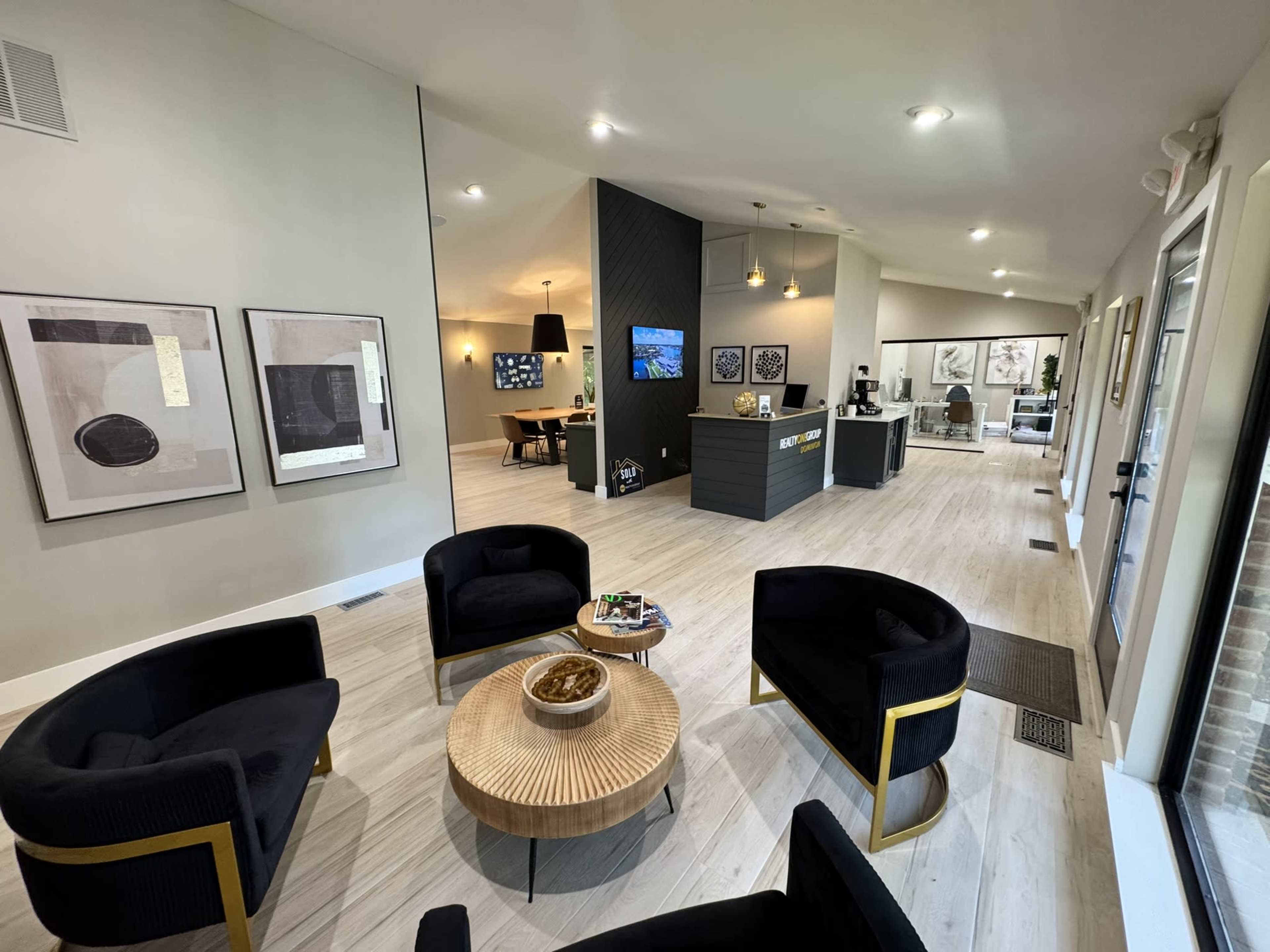 The image shows a modern reception area with black chairs arranged around a circular wooden table, artworks on the wall, and a front desk in the background.