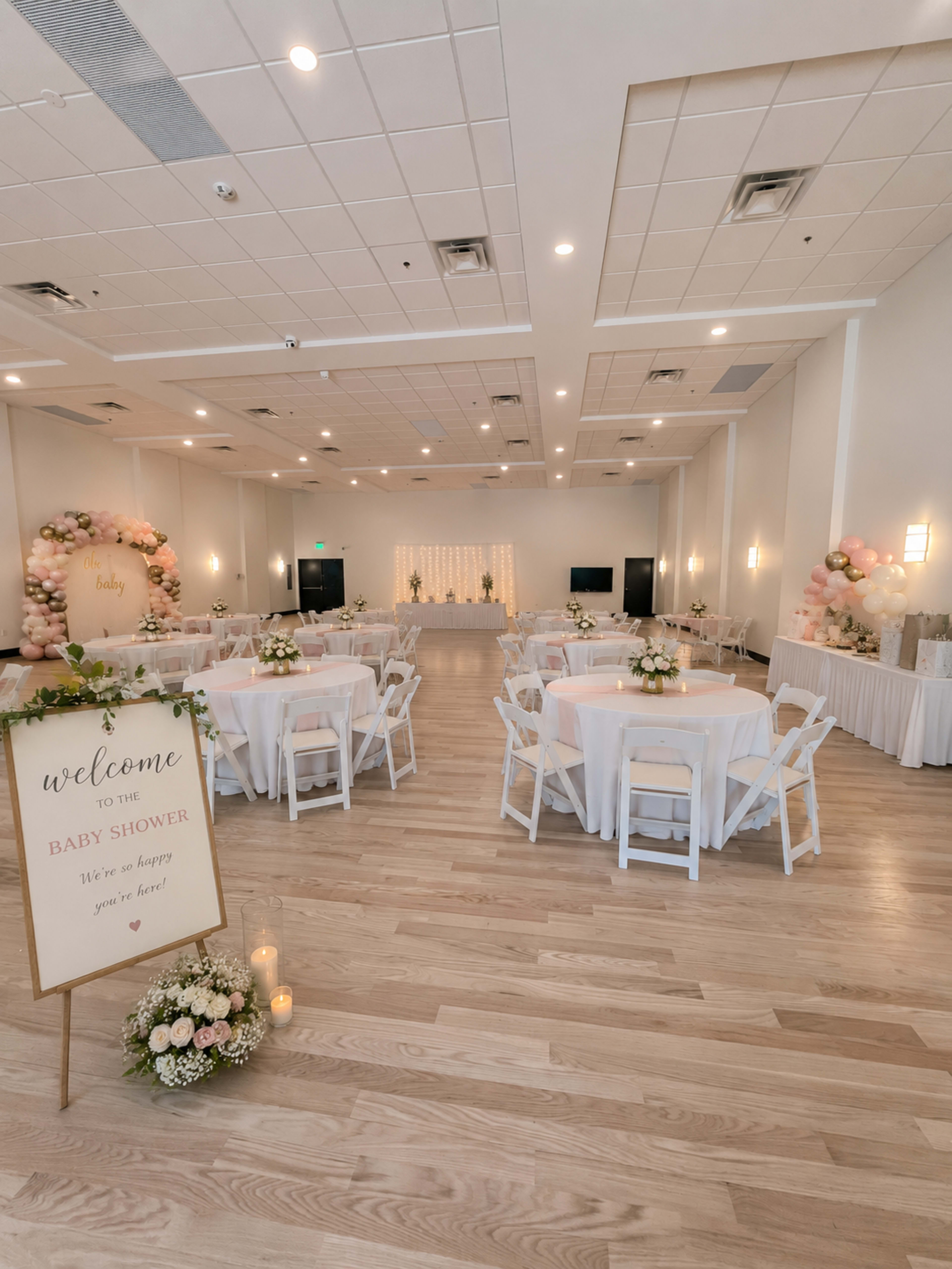 The image shows a decorated event space for a baby shower, featuring round tables with white tablecloths, floral centerpieces, and a welcome sign at the entrance.