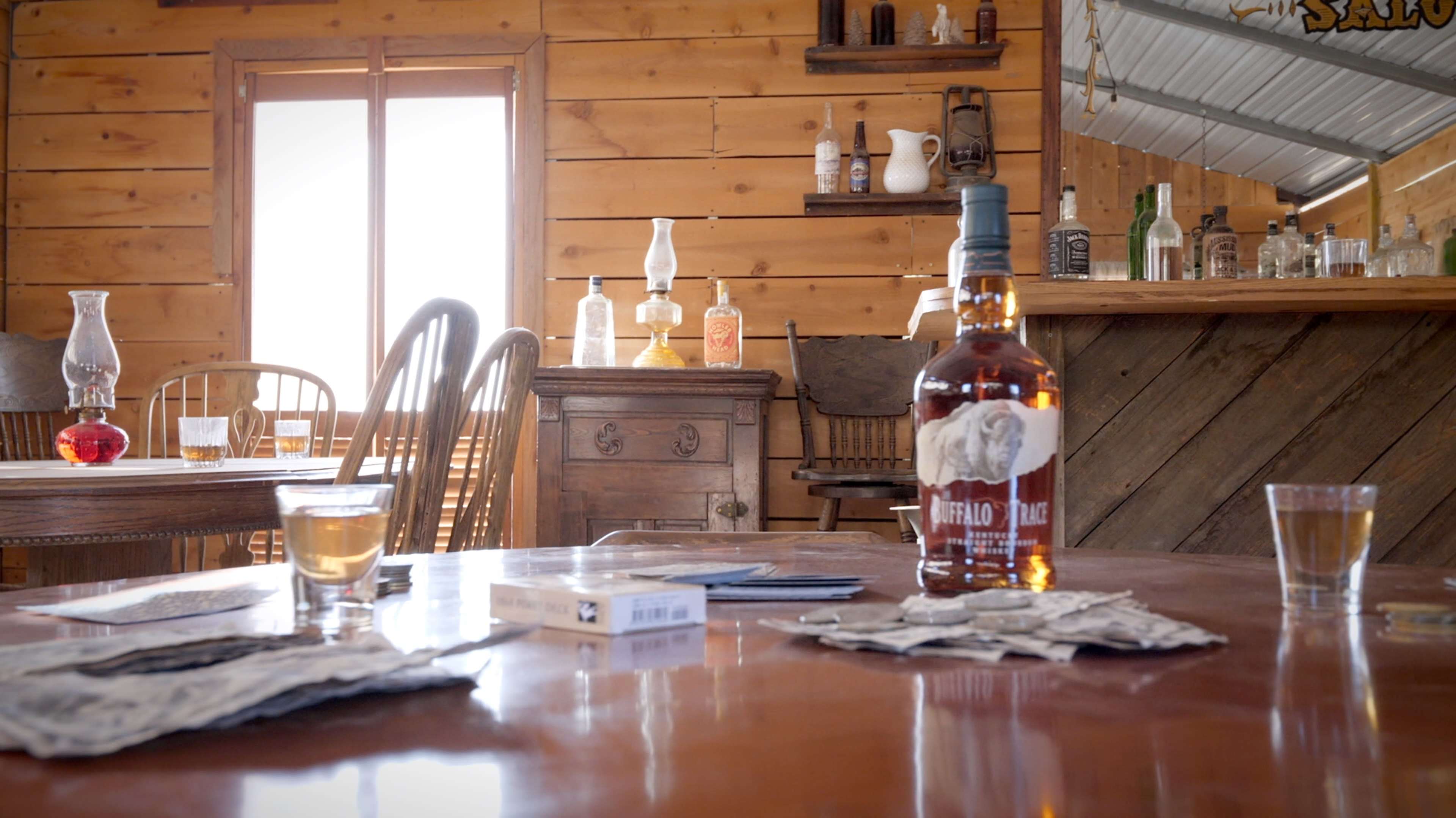 A rustic interior with a wooden table featuring a bottle of Buffalo Trace whiskey and shot glasses, alongside stacks of playing cards, set against a backdrop of shelves displaying vintage bottles and a window reflecting natural light.
