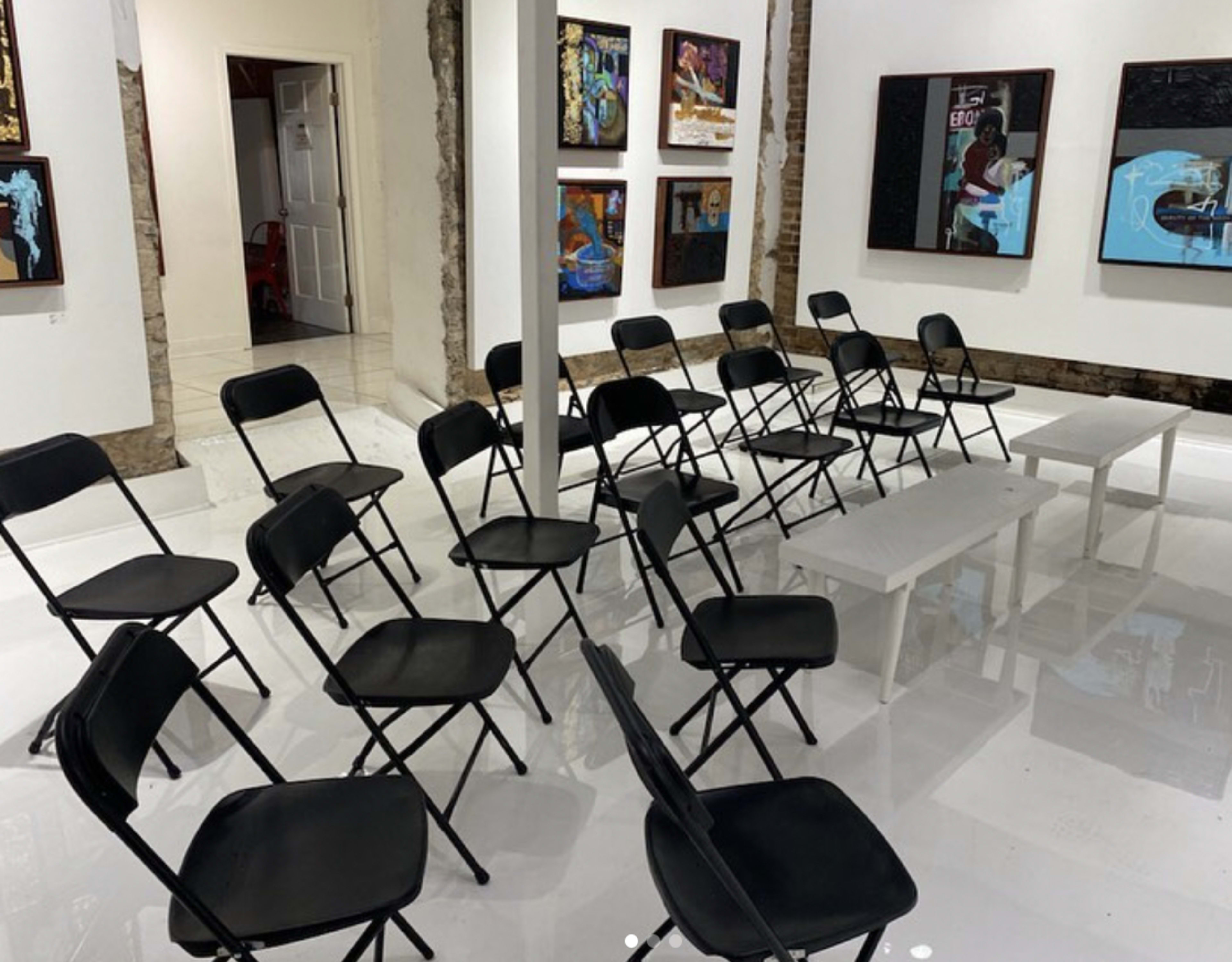 A room with several black folding chairs arranged in a circular pattern around a small white table, with artwork displayed on the walls.