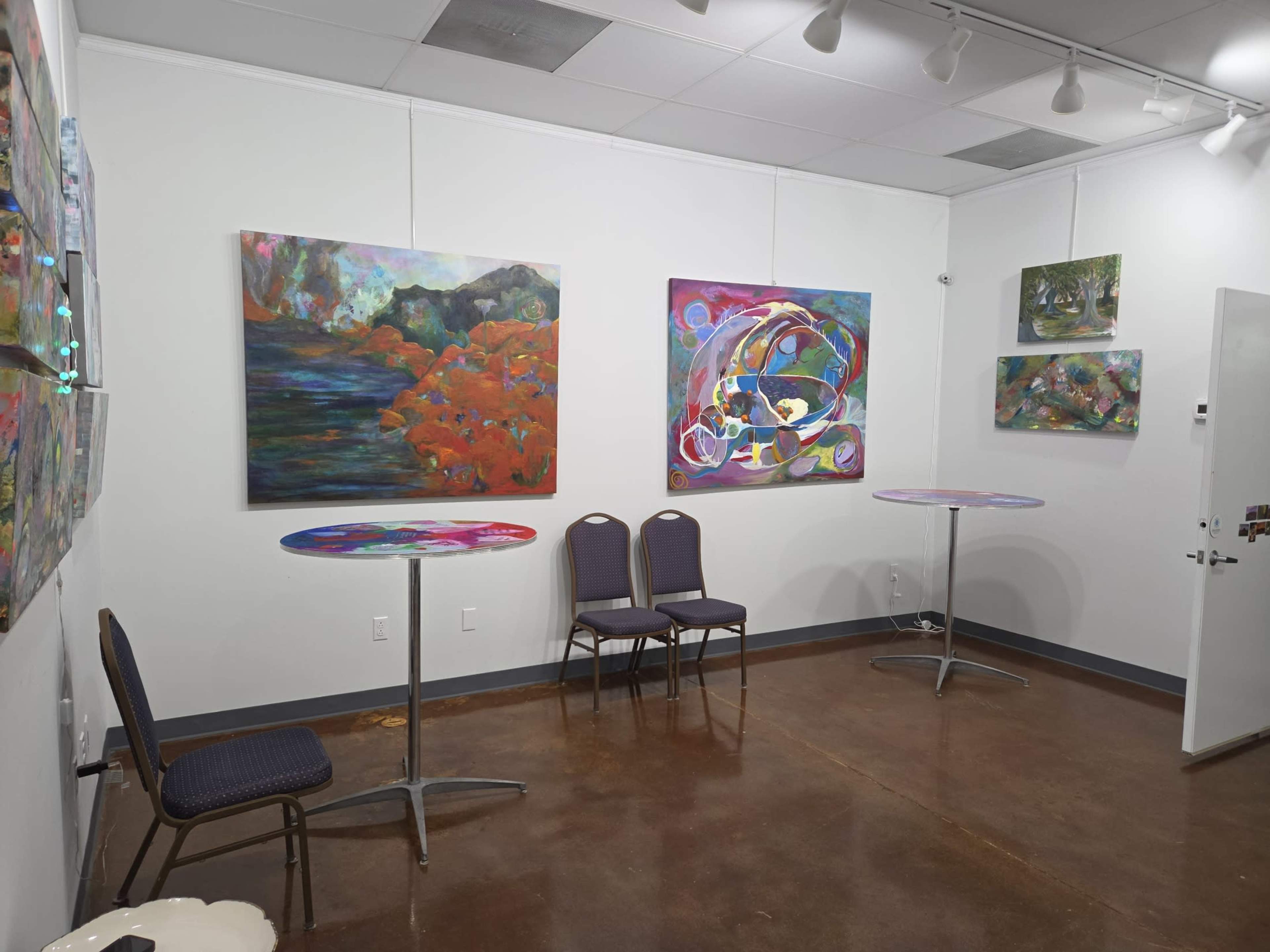 The image shows an art gallery interior featuring colorful paintings on the walls, two high tables, and two chairs in a modern setup.