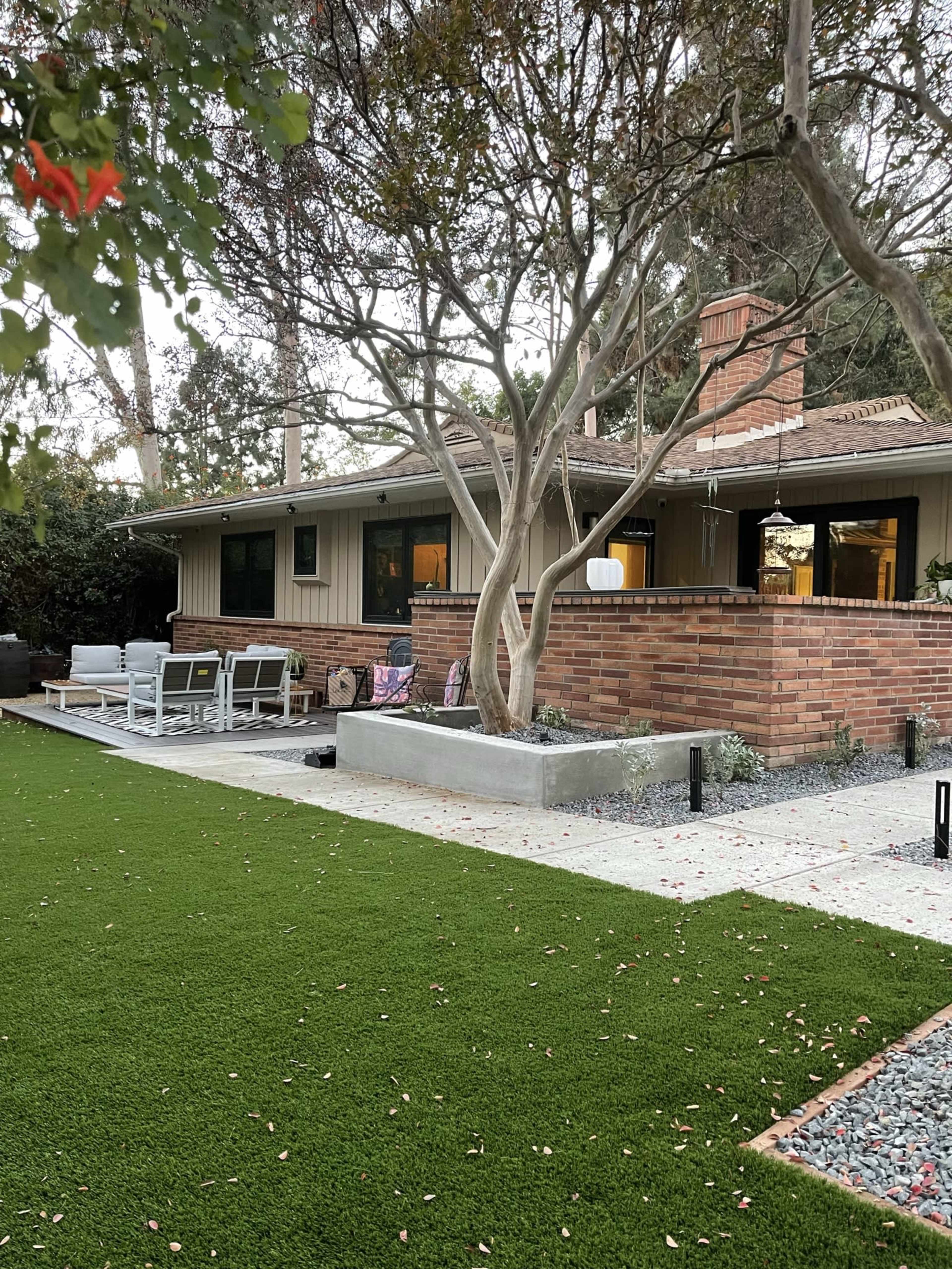 A house with brick accents and a patio area is surrounded by a manicured lawn and trees.