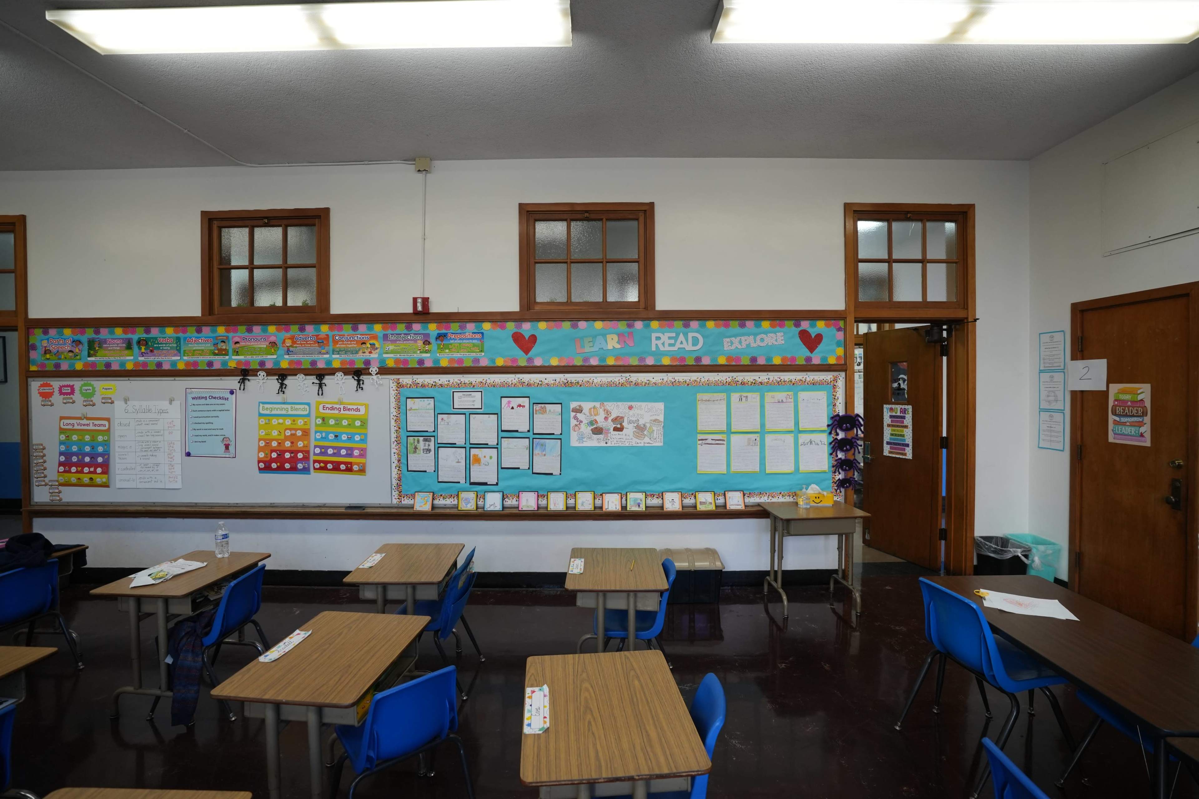 The image shows a classroom with multiple tables and blue chairs, along with a bulletin board displaying educational materials and announcements on the walls.