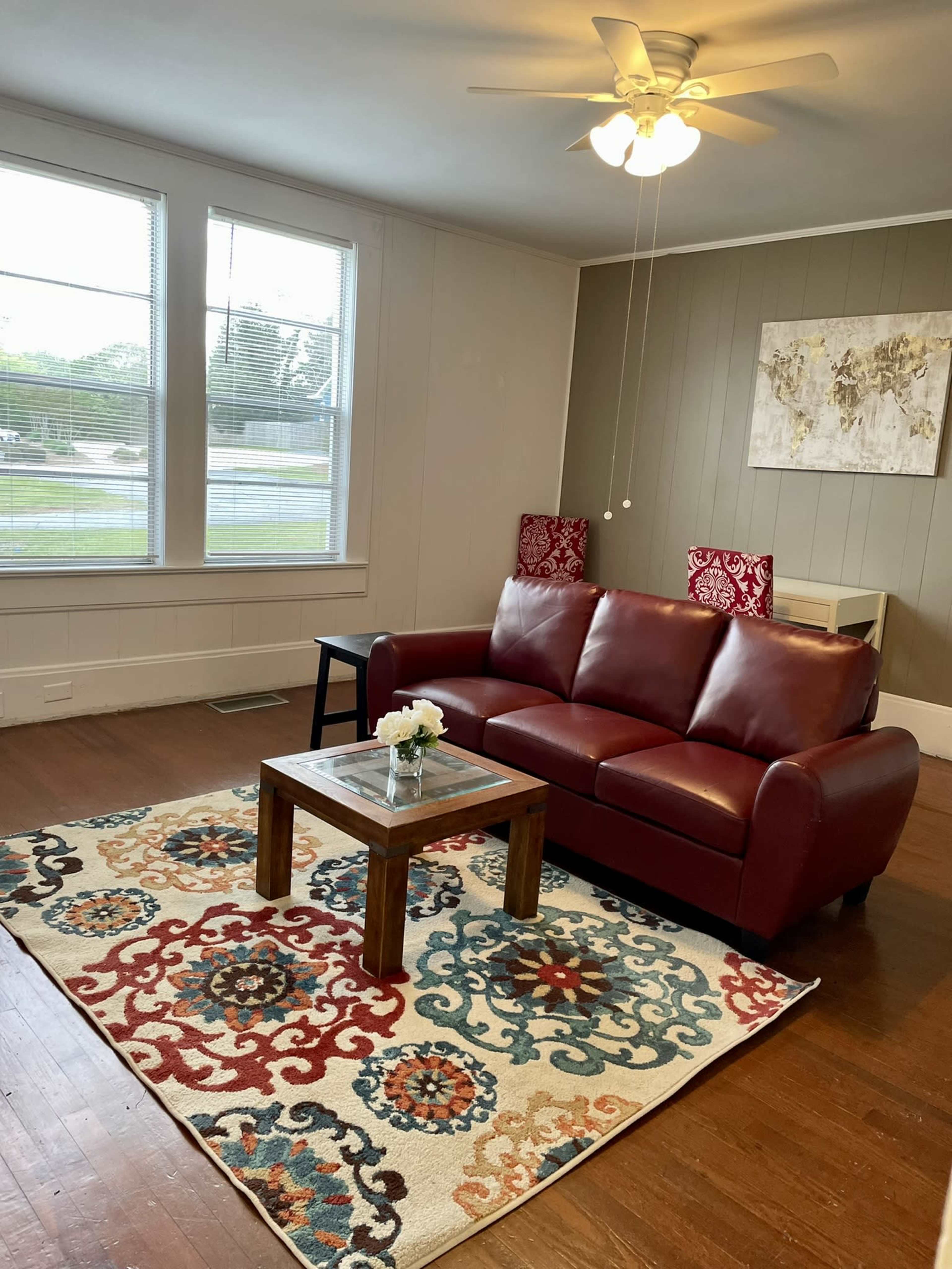 A red sofa is placed on a patterned rug in a living room with large windows and a ceiling fan.