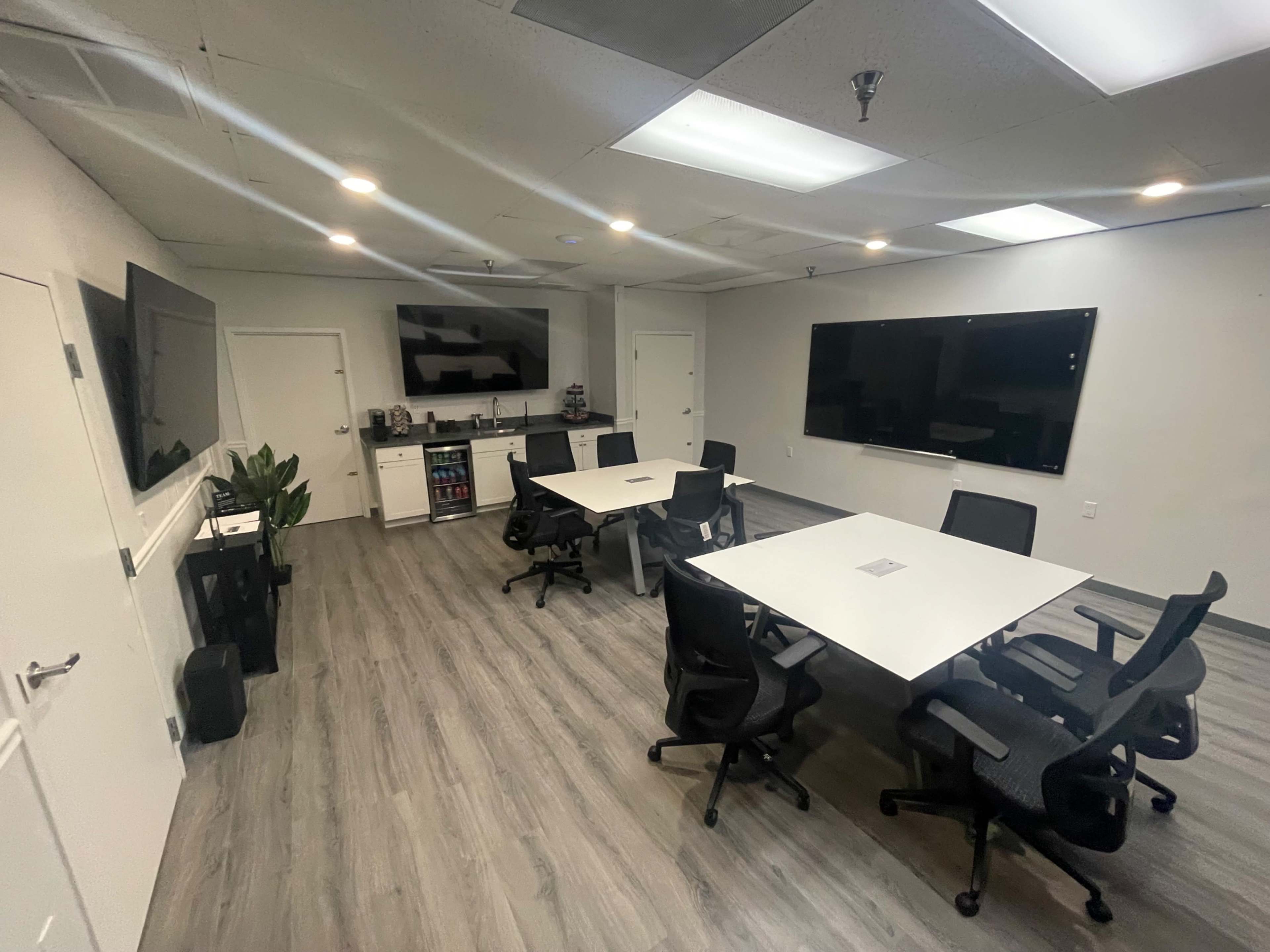 A modern office meeting room with several white tables and black chairs, featuring a kitchenette area in the background.