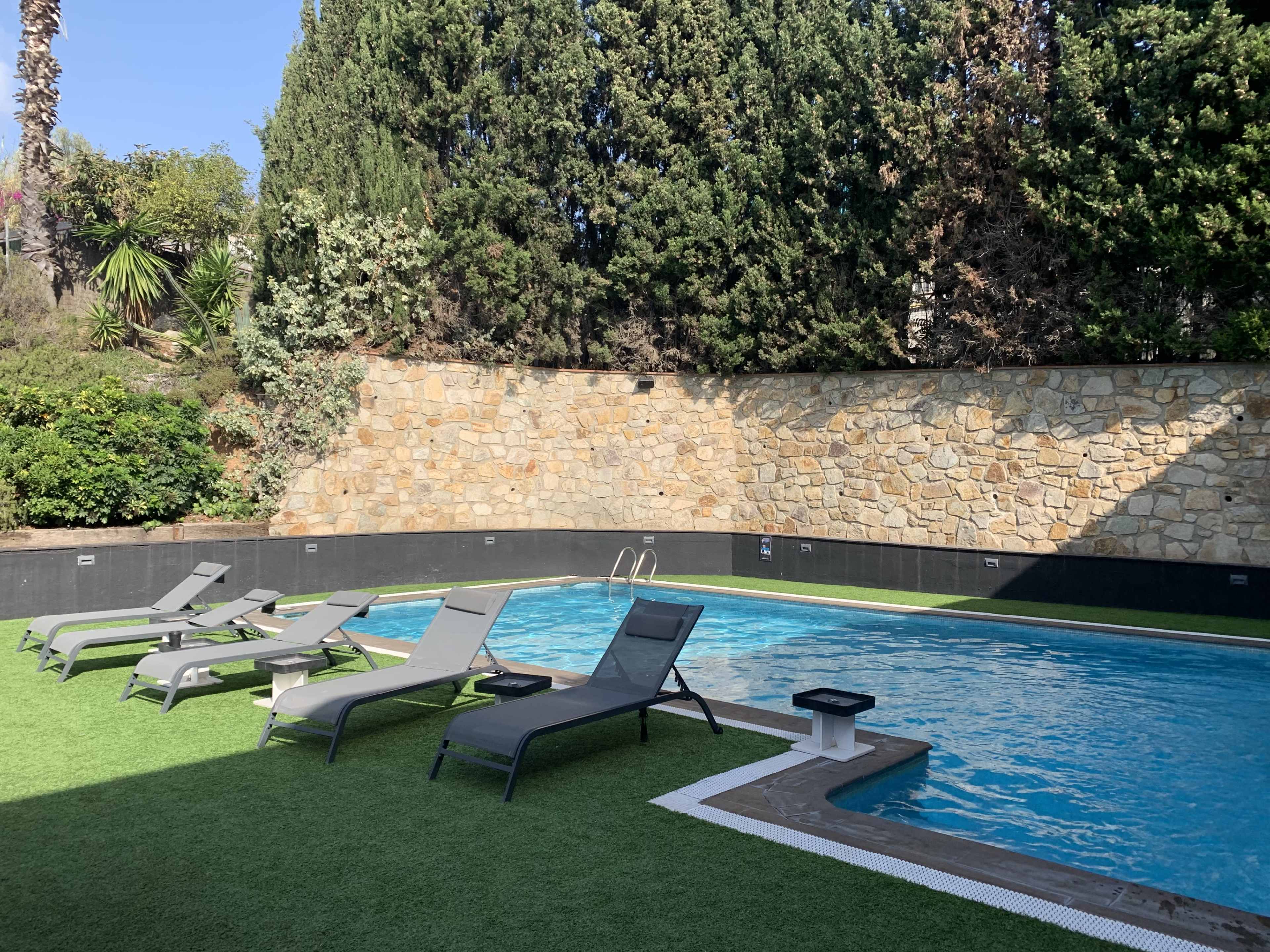 The image shows a swimming pool surrounded by lounge chairs on a grassy area, enclosed by a stone wall and greenery.