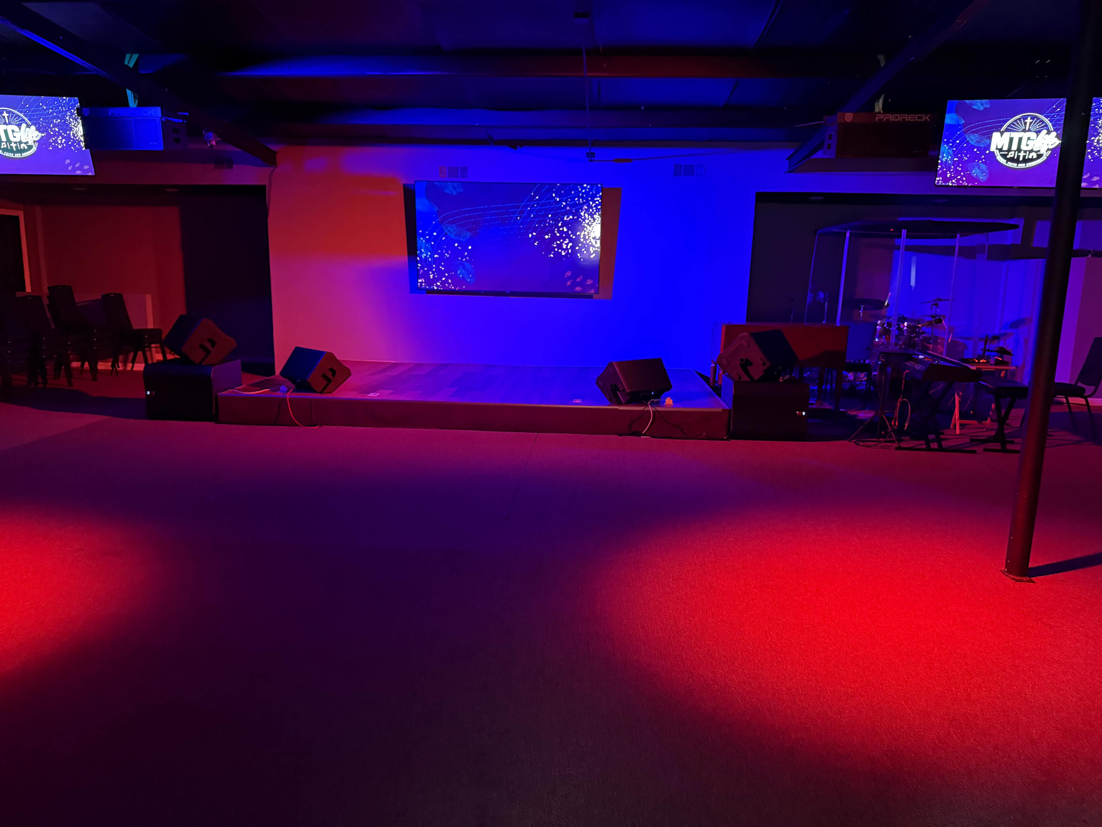 The image shows a performance space with a large screen, sound equipment, and red and blue lighting.