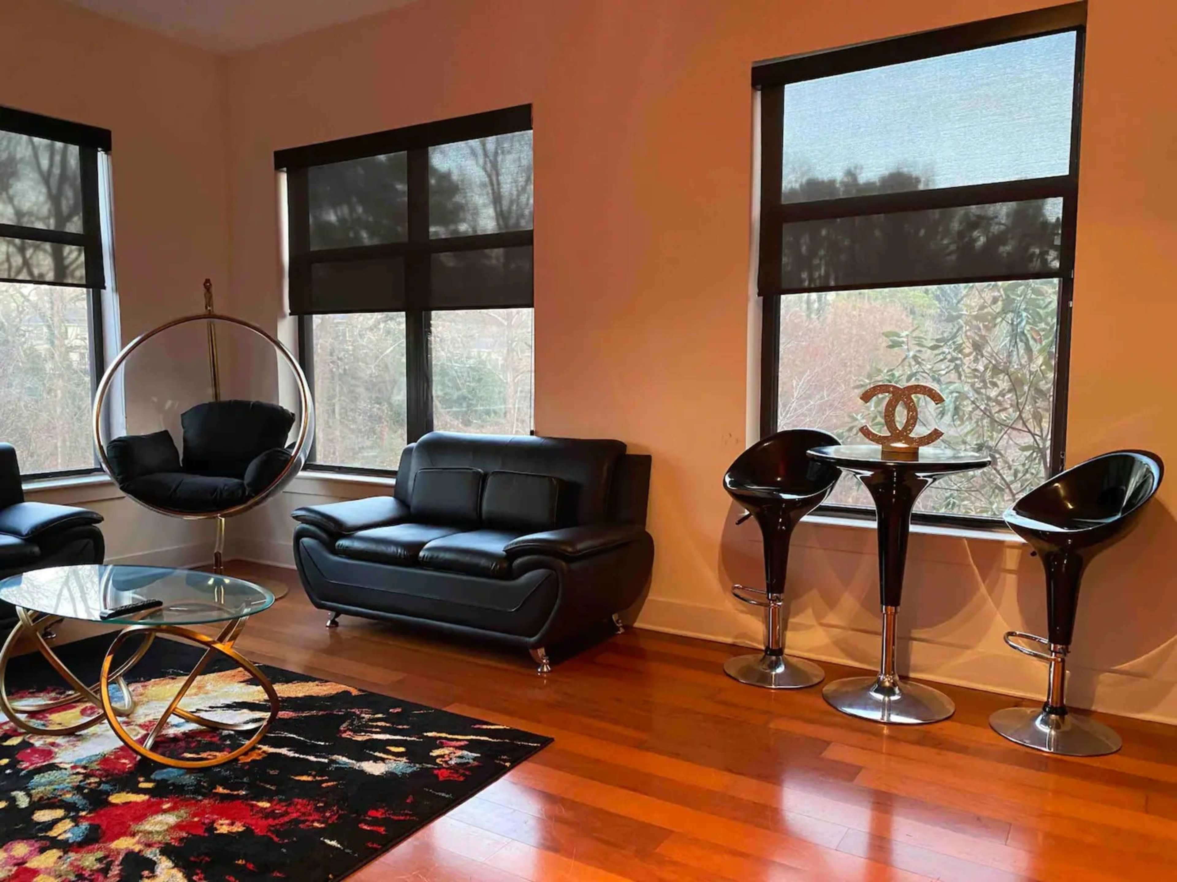 The room features a black leather sofa, a circular hanging chair, and a glass coffee table on a colorful rug, with two sleek bar stools beside a round table.