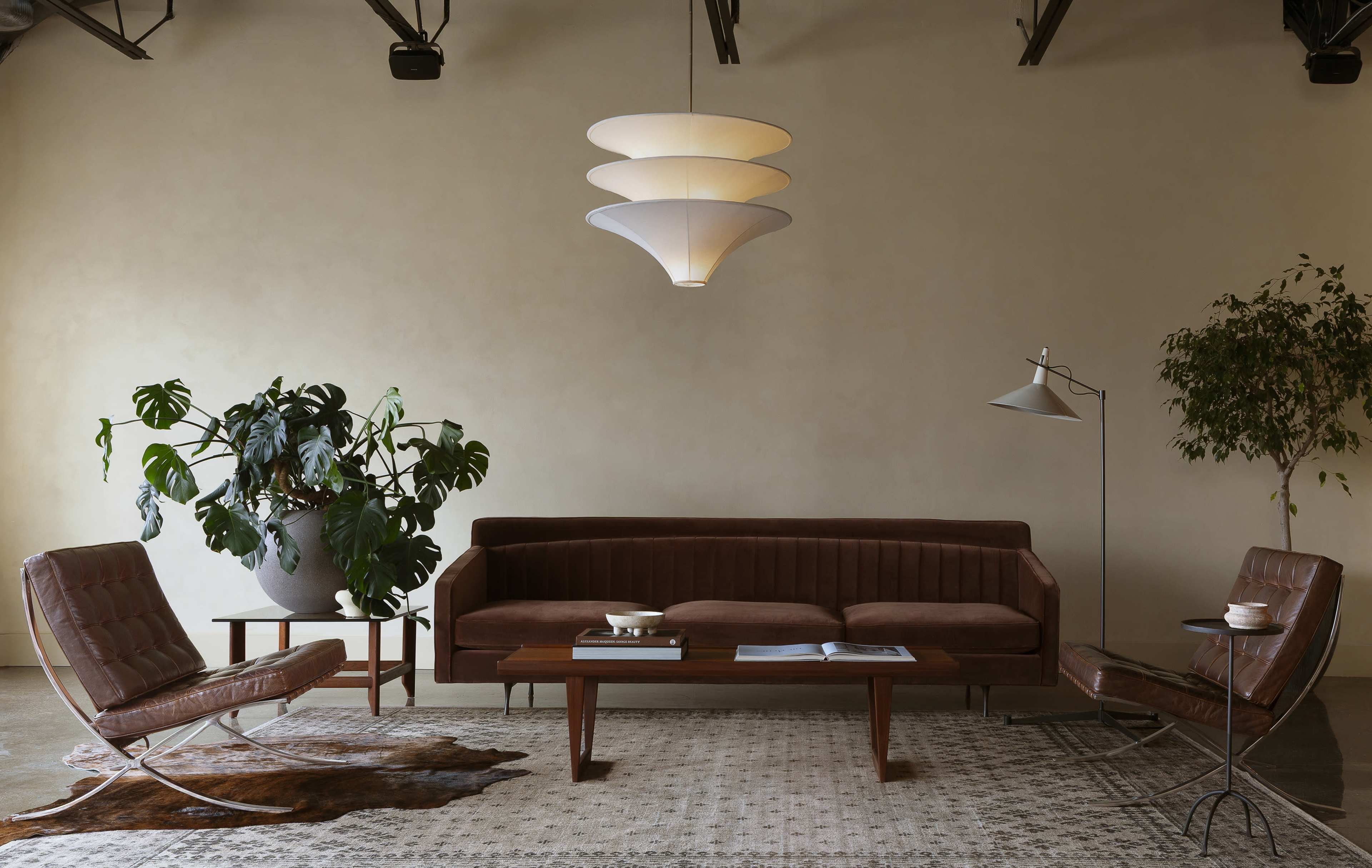 The scene features a minimalist living room with a brown sofa, two stylish chairs, a wooden coffee table, and a large potted plant, all set against a neutral wall.