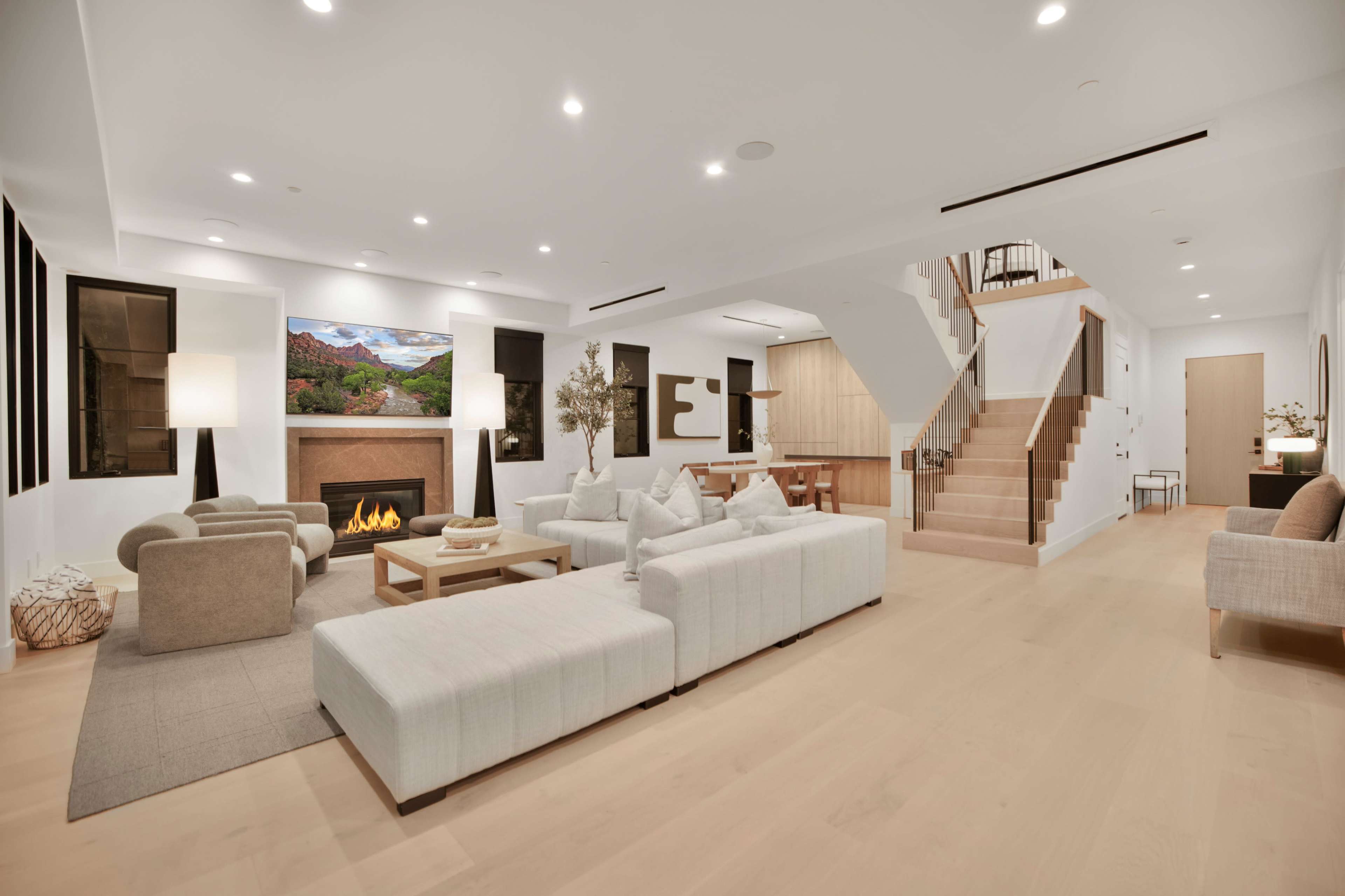 The image shows a spacious living room featuring a modern design with a fireplace, a large sectional sofa, a dining area, and stairs leading to an upper level.