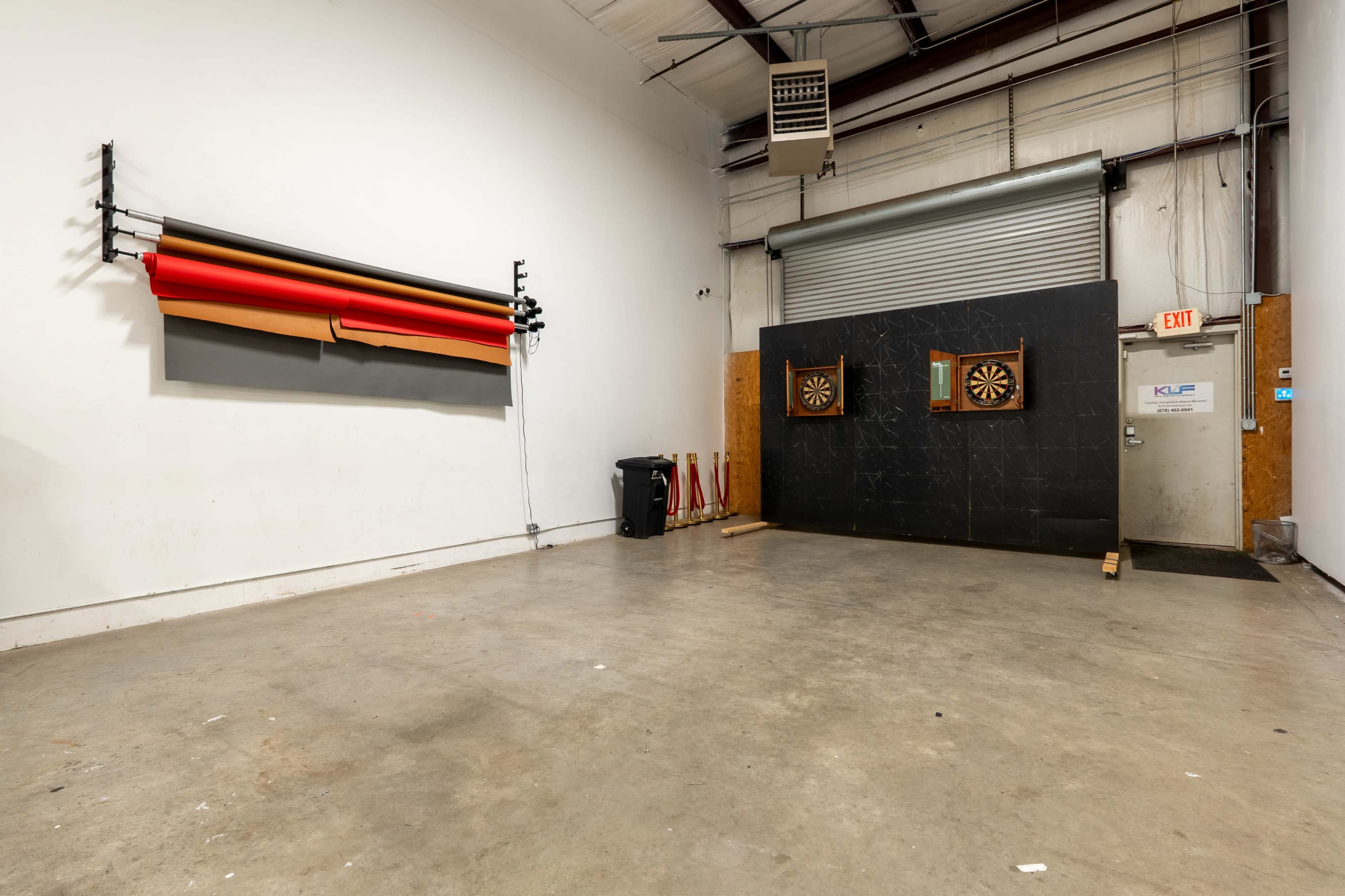The image shows a spacious room with a set of dartboards mounted on a wall and a roll of colored paper hanging on the opposite side.