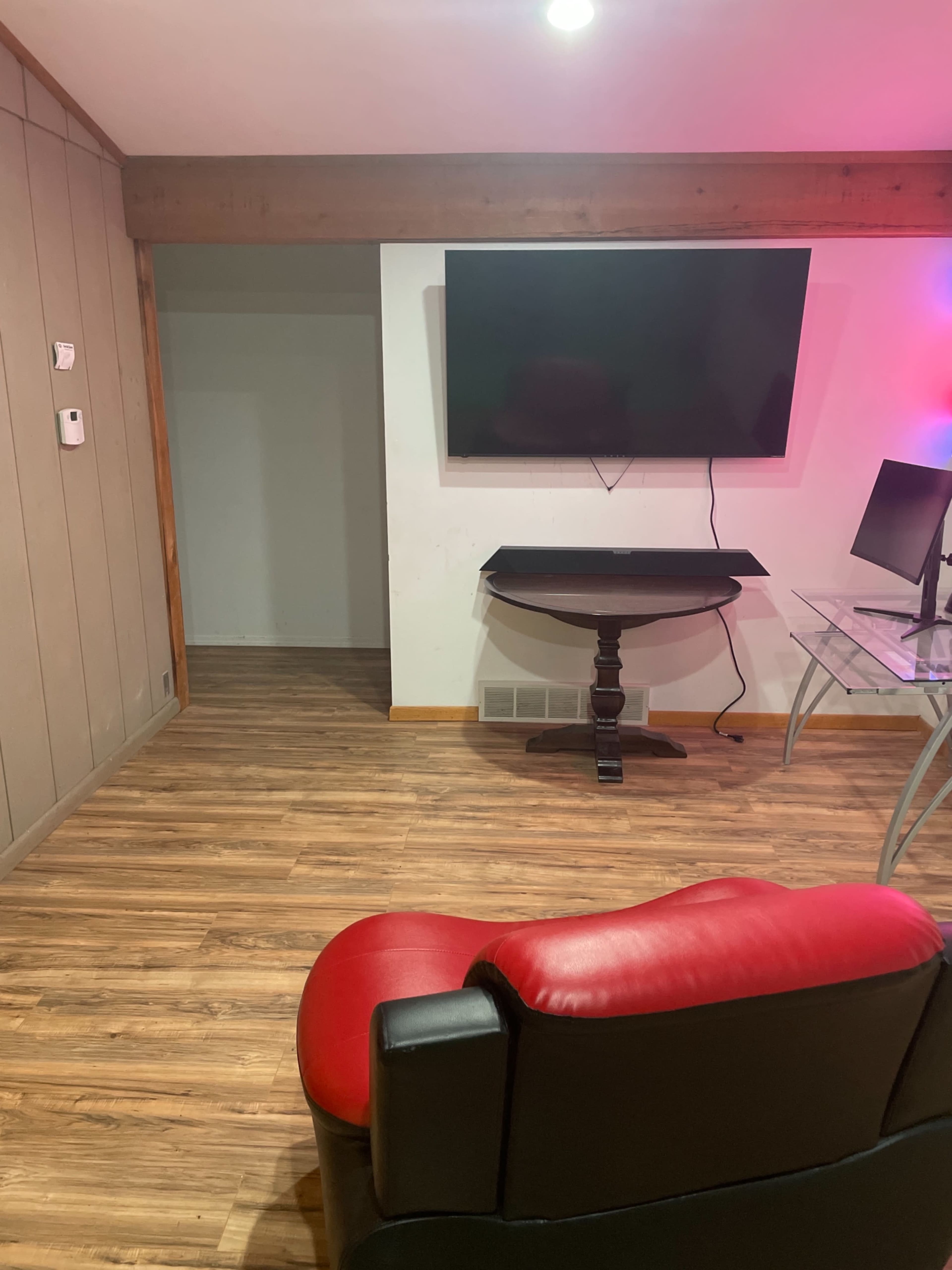A room with a hardwood floor, a television mounted on the wall, a small table in front, and a red chair positioned to the side.