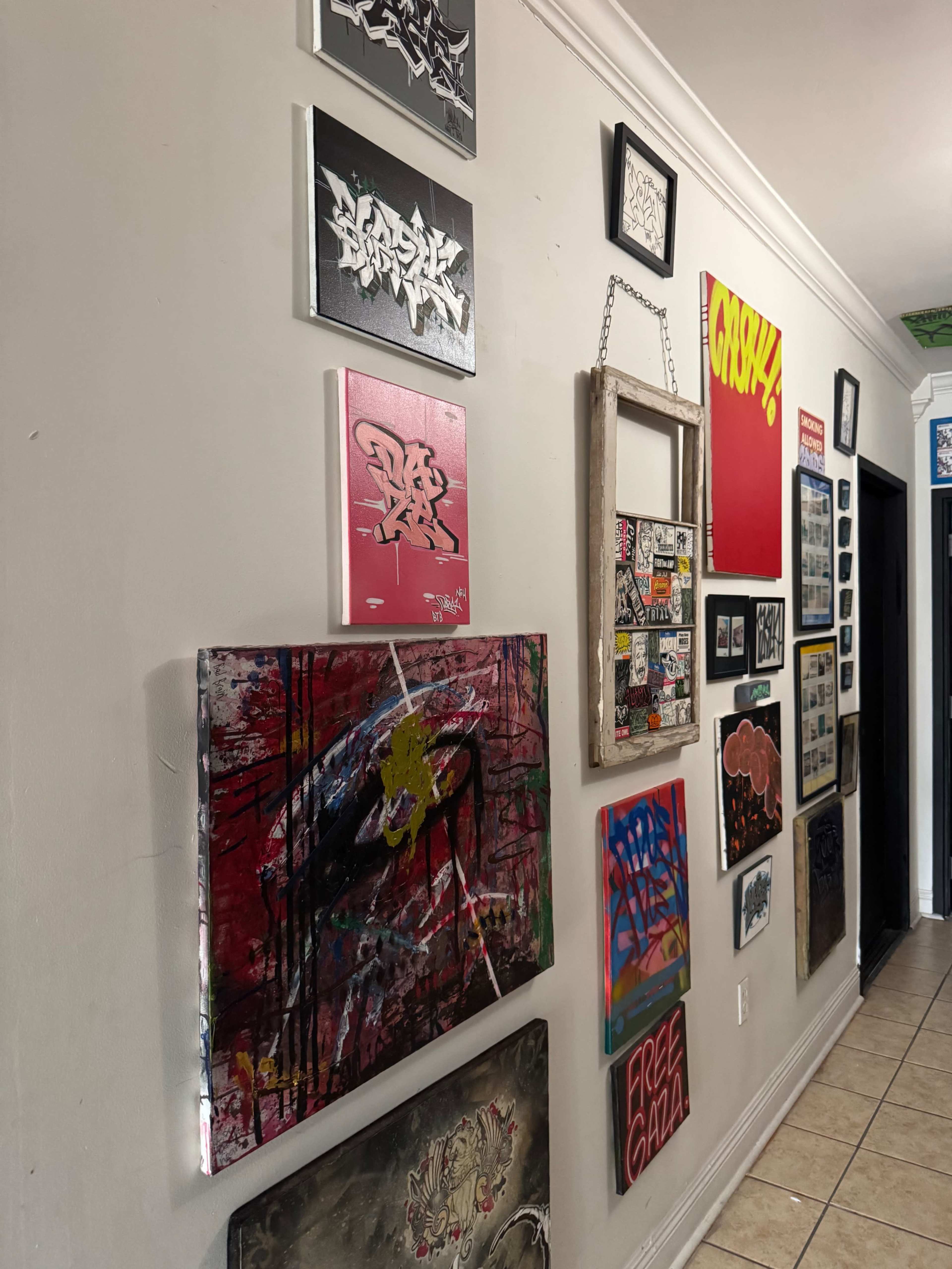 The image shows a wall adorned with various pieces of graffiti art and framed paintings in a hallway setting.