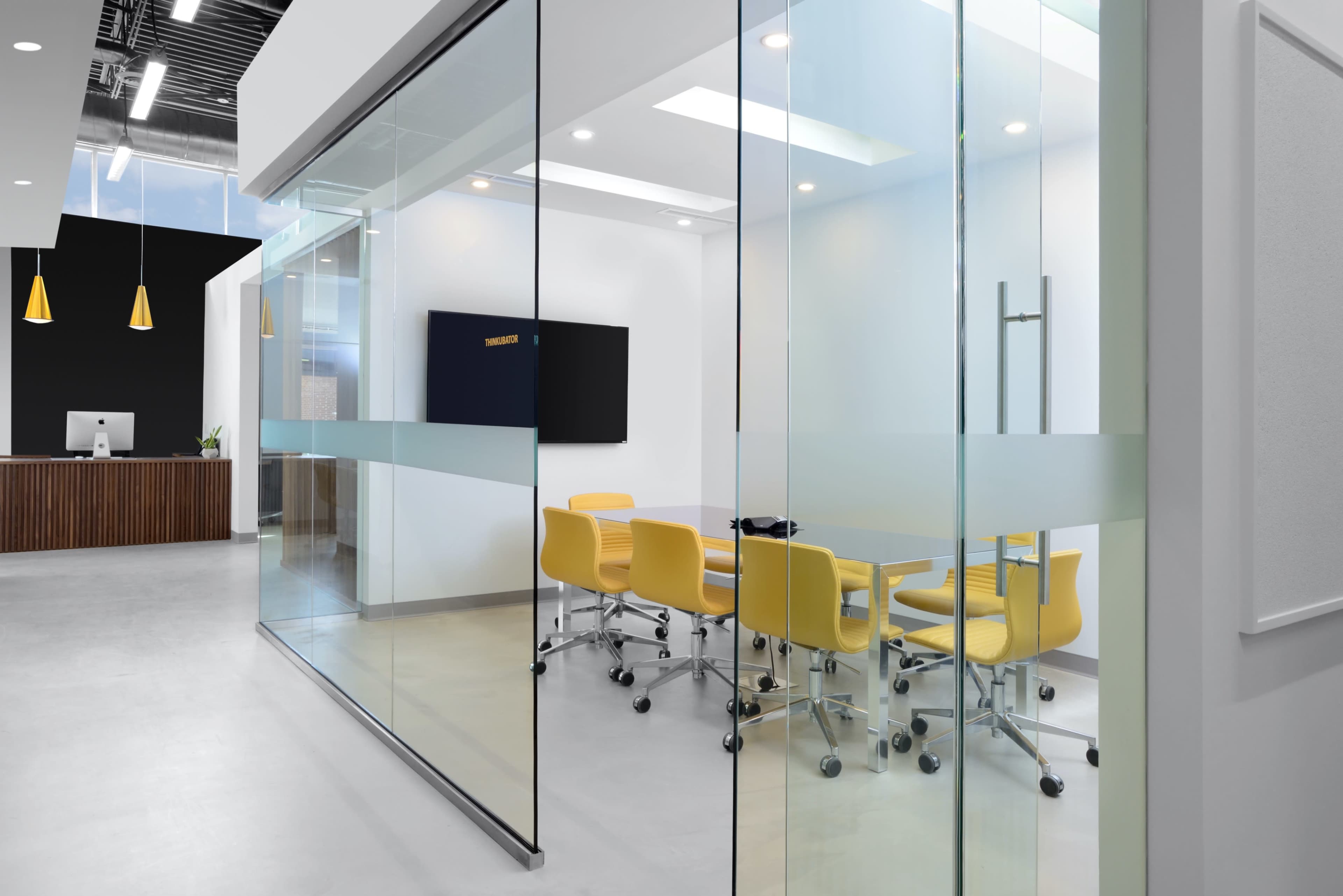 A modern glass-walled conference room features yellow chairs and a large screen, adjacent to a reception area with a wooden counter.