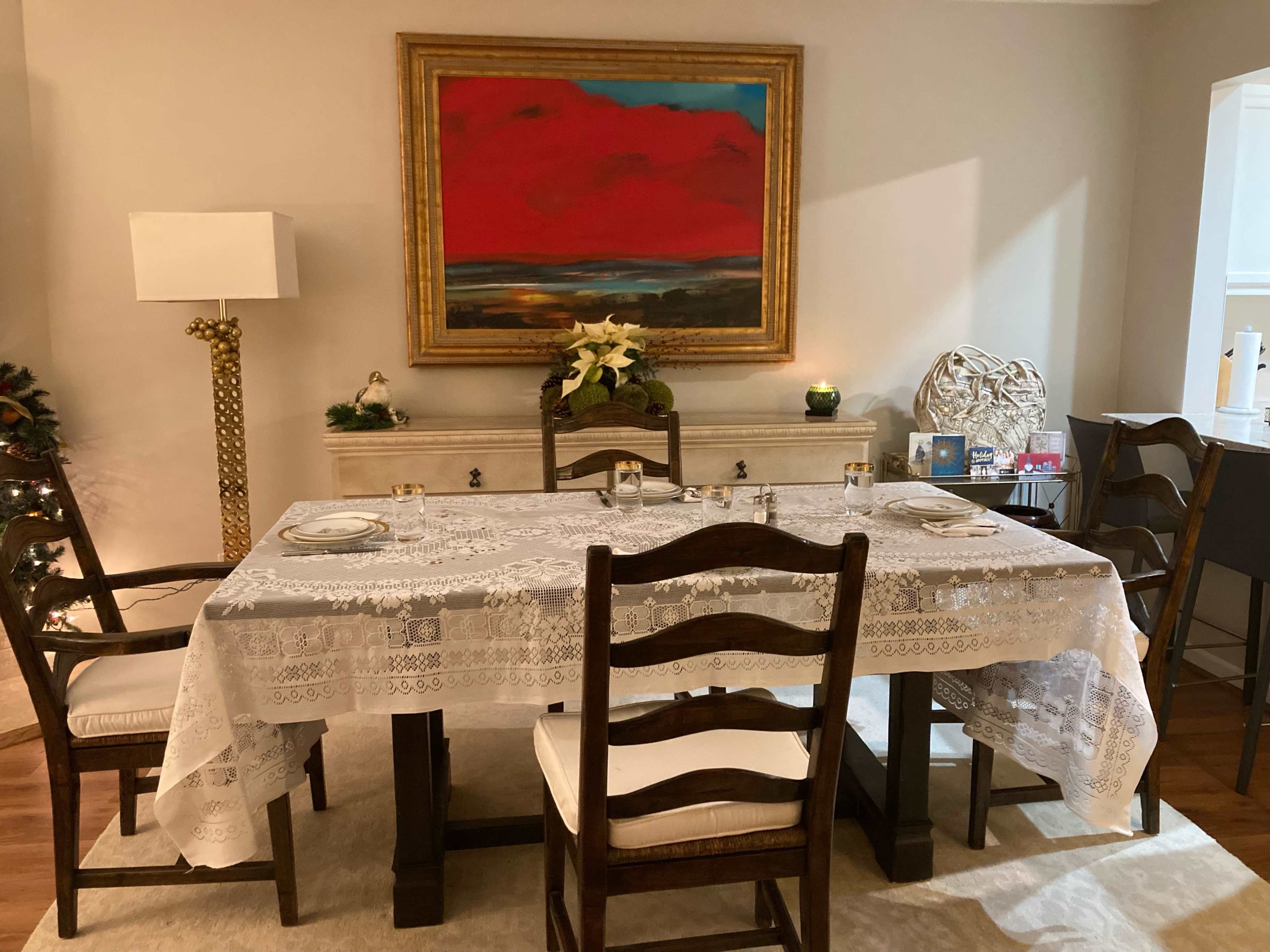 A dining table set with a lace tablecloth and four wooden chairs is positioned in a room with a large painting and a decorative lamp nearby.