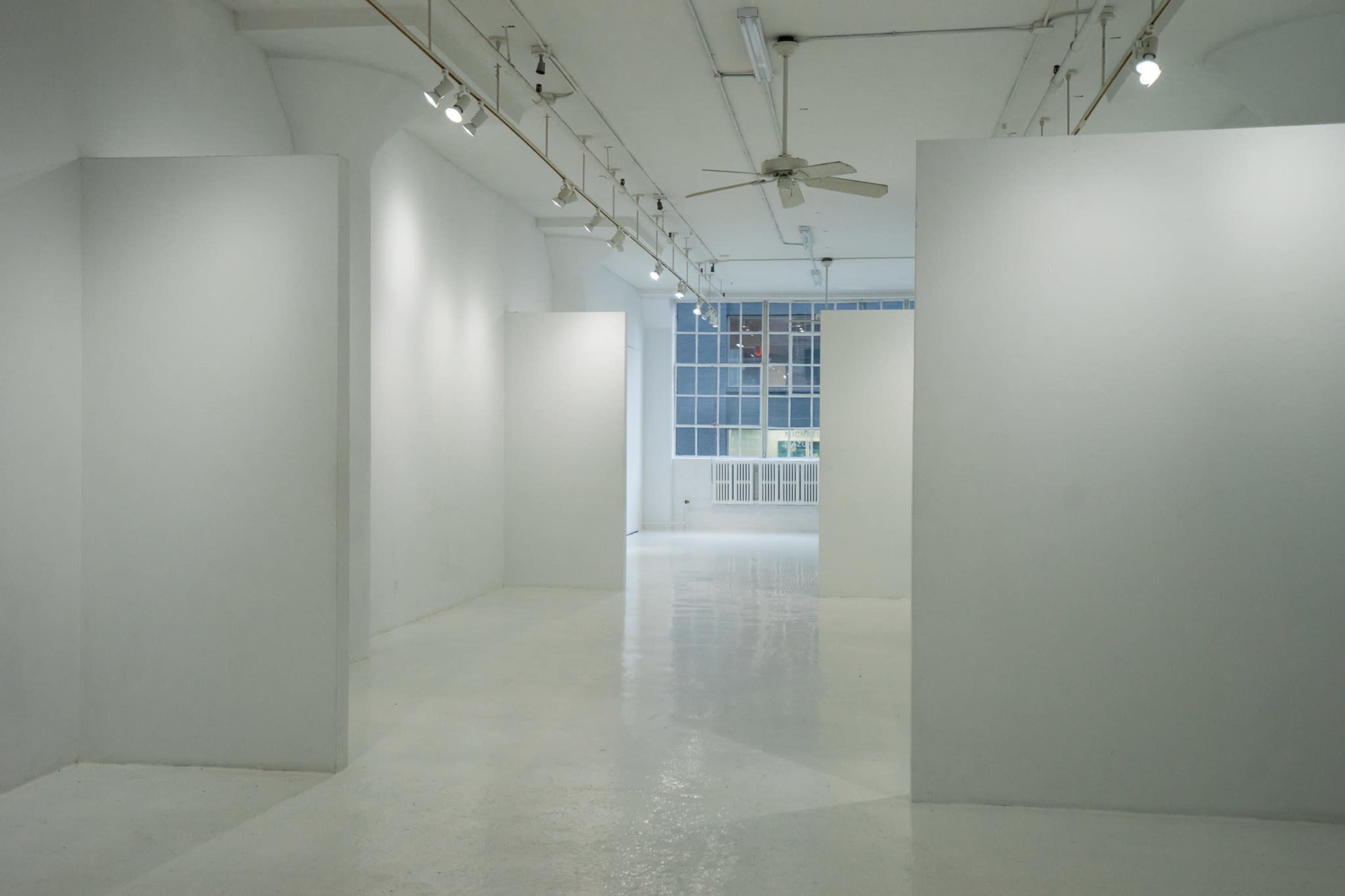 The image shows a spacious, empty art gallery featuring white walls and floors, with bright lighting and large windows in the background.