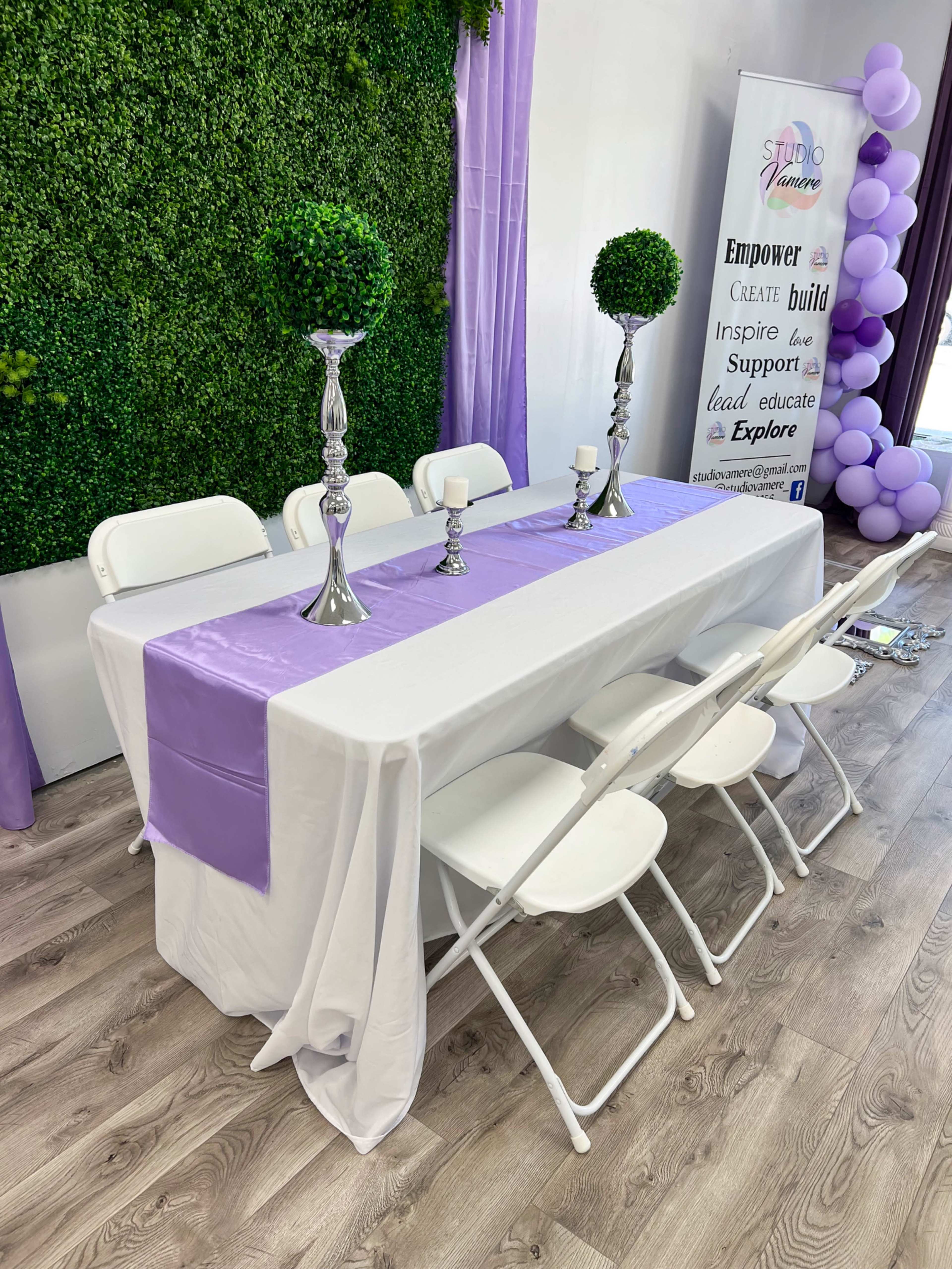 A dining table is set with a white tablecloth, a lavender table runner, silver candlesticks, and decorative green topiaries against a backdrop of greenery and balloons.