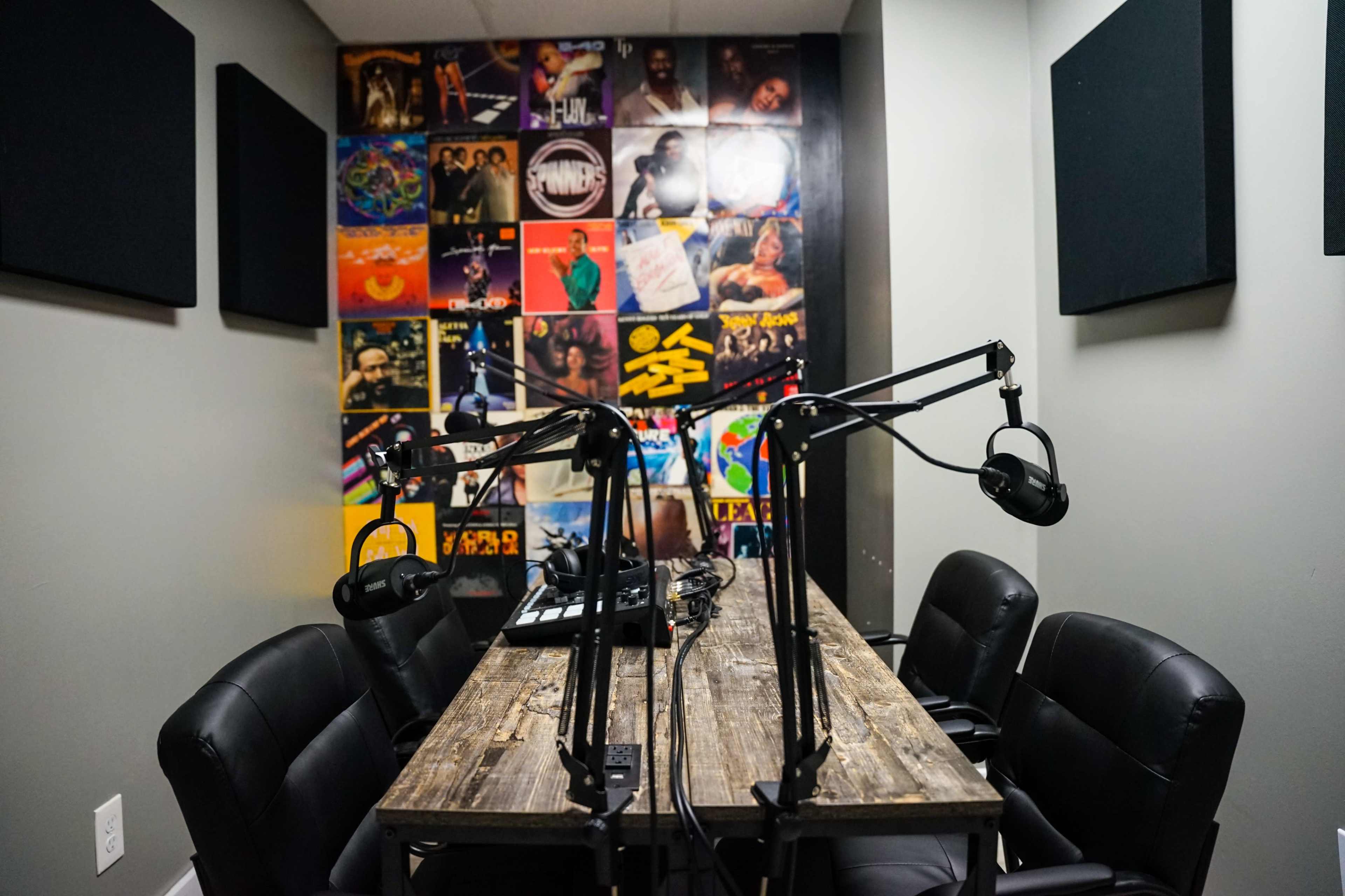 A small recording studio features a wooden table surrounded by black chairs and two microphone setups, with a wall covered in colorful album art.
