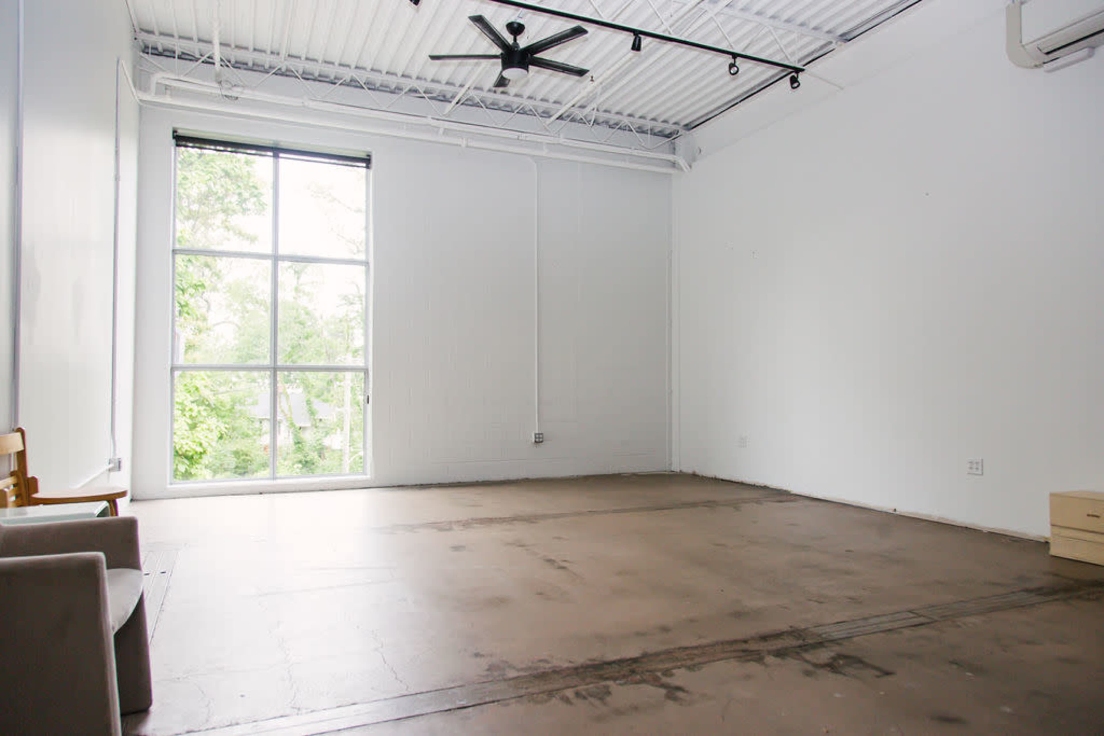 A spacious, empty room with a large window overlooking greenery, a ceiling fan, and minimal furnishings.