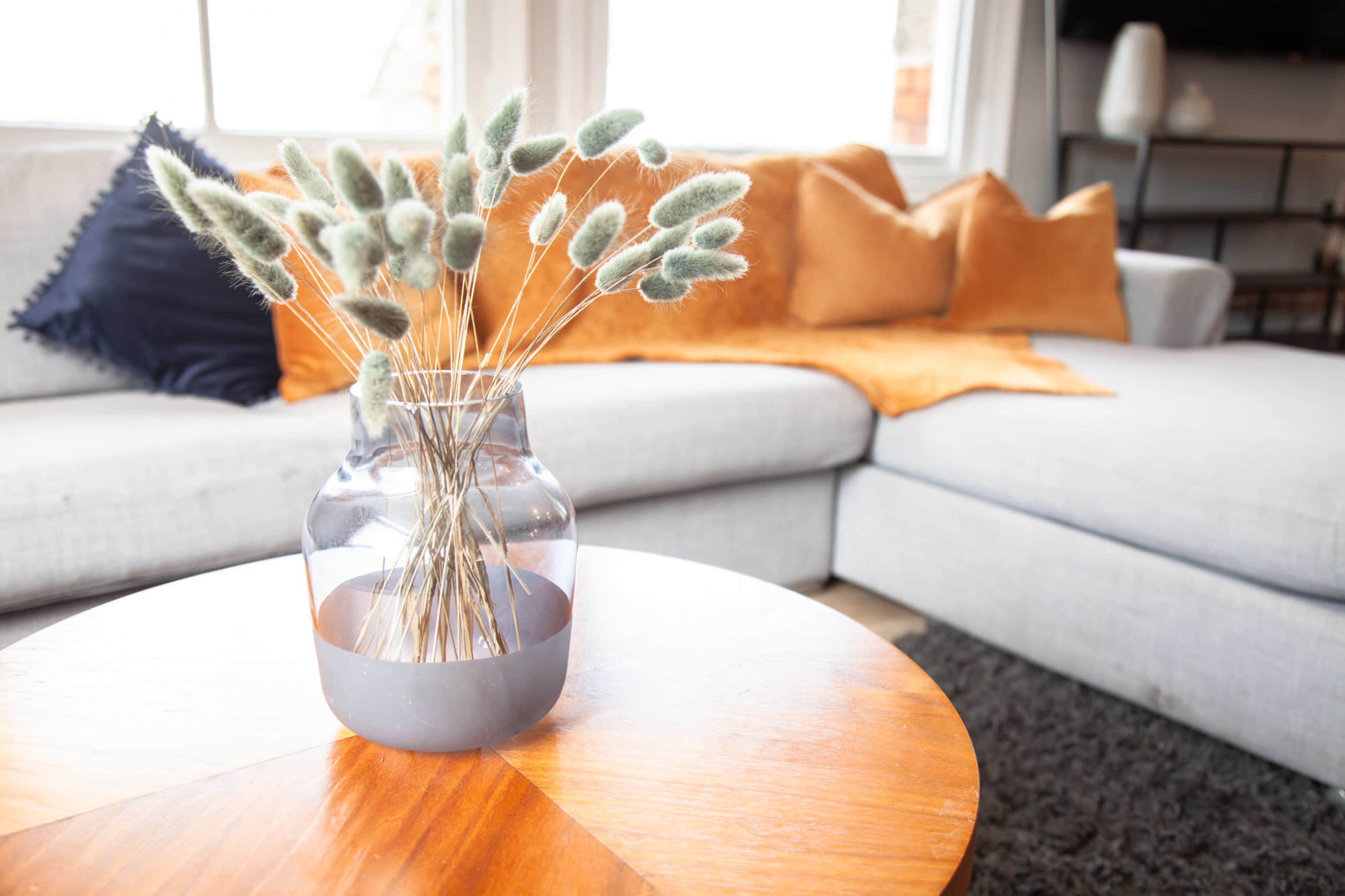 A clear vase filled with decorative dried plants sits on a wooden coffee table in a living room featuring a gray sectional sofa and orange throw pillows.