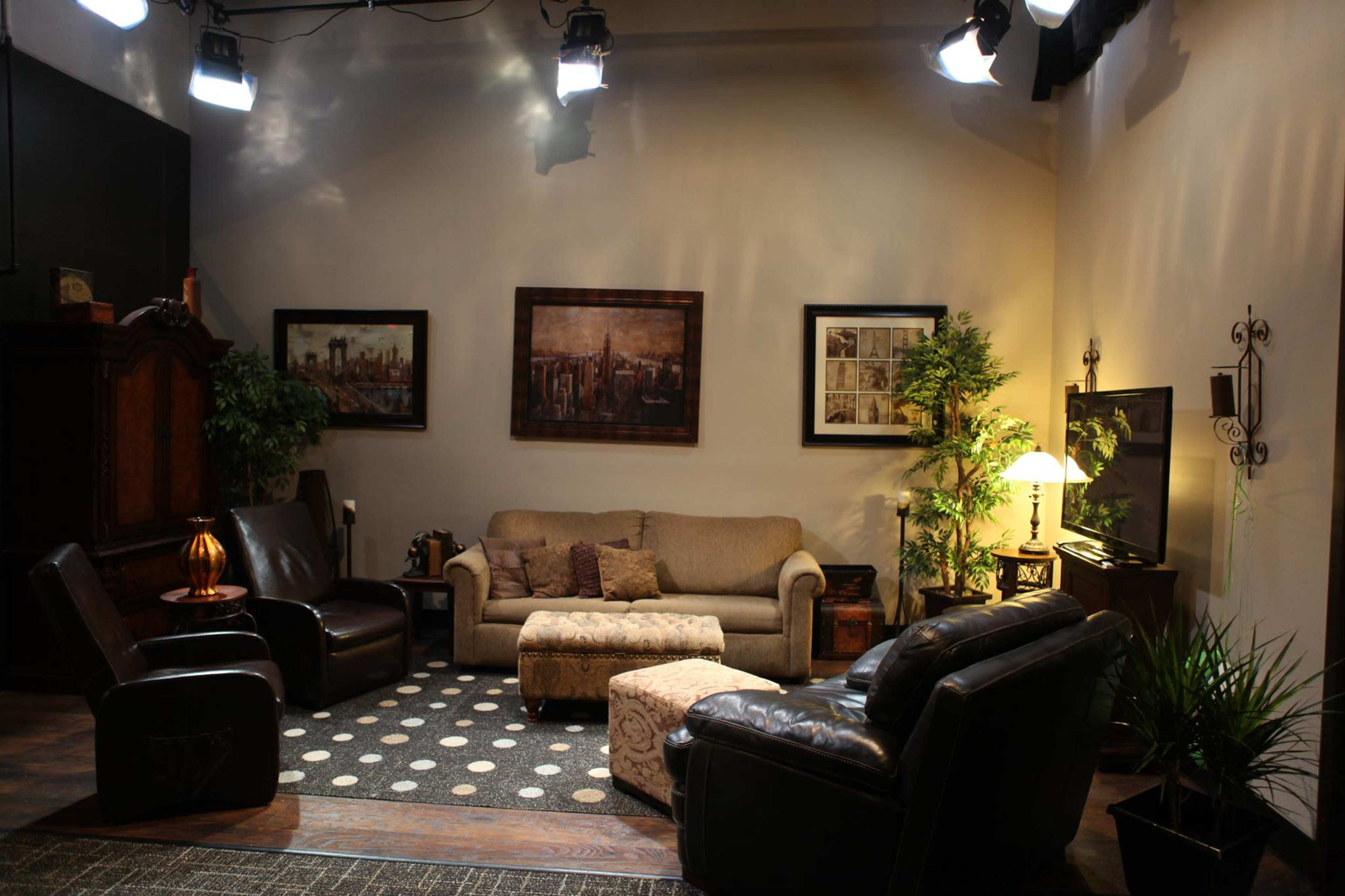 The image shows a cozy living room with a sofa, two armchairs, a coffee table, and decorative plants, all under bright studio lights.