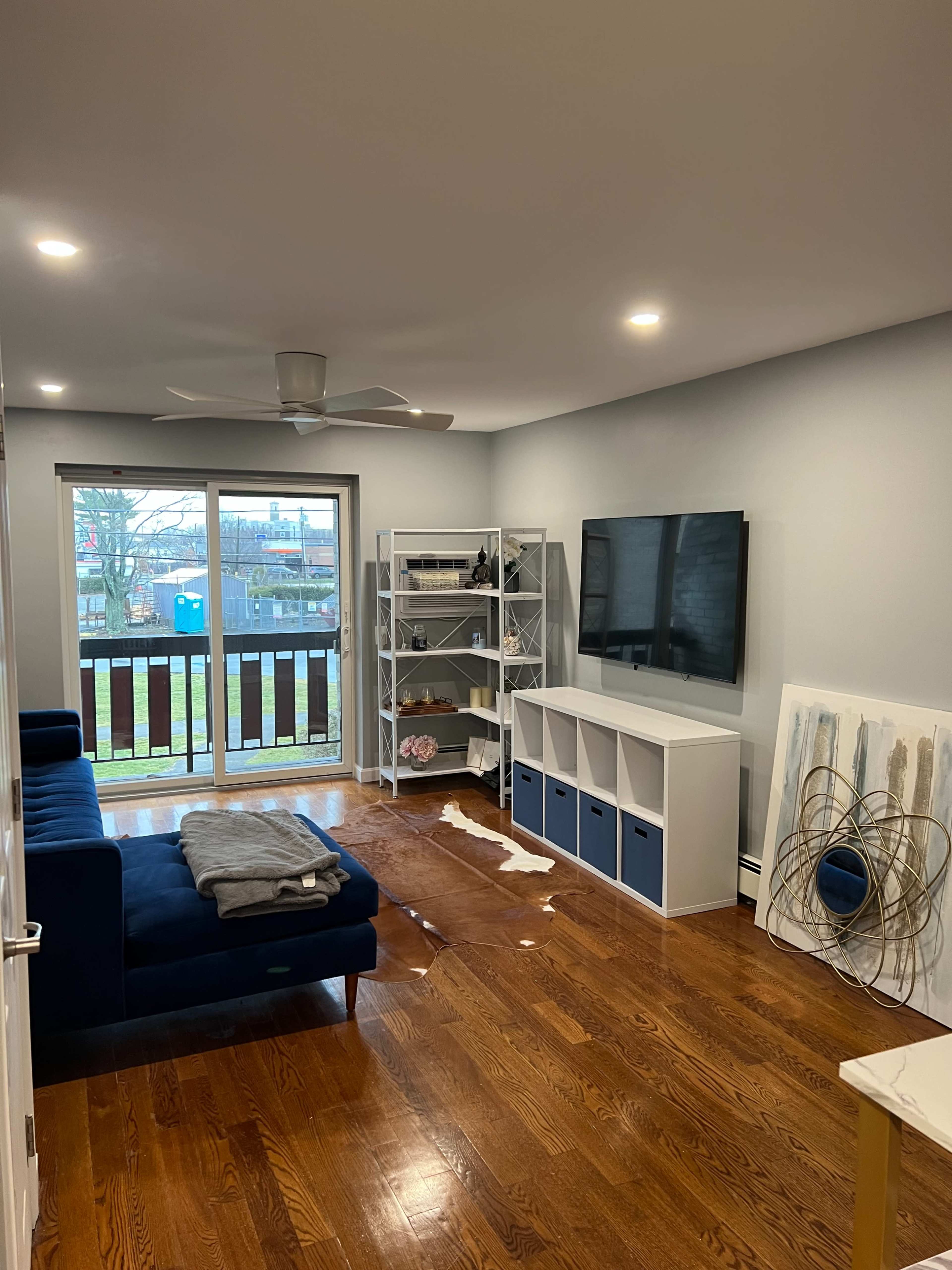 A living room features a dark blue sofa, a TV mounted on the wall, and a bookshelf filled with decorative items, next to sliding glass doors leading to a balcony.