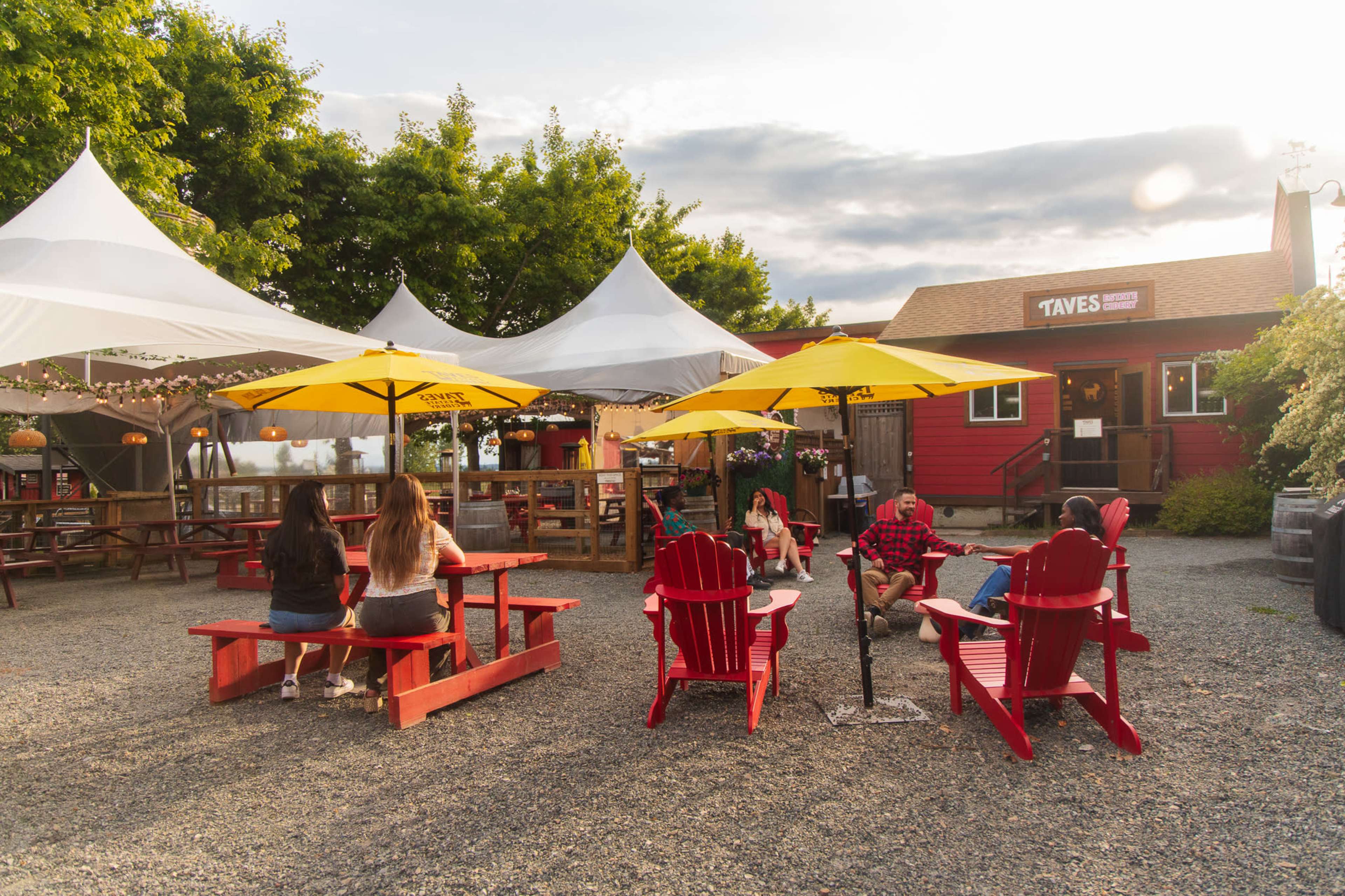 A group of people sits at picnic tables under yellow umbrellas in a gravel courtyard beside a red building with a sign that reads "Taves."