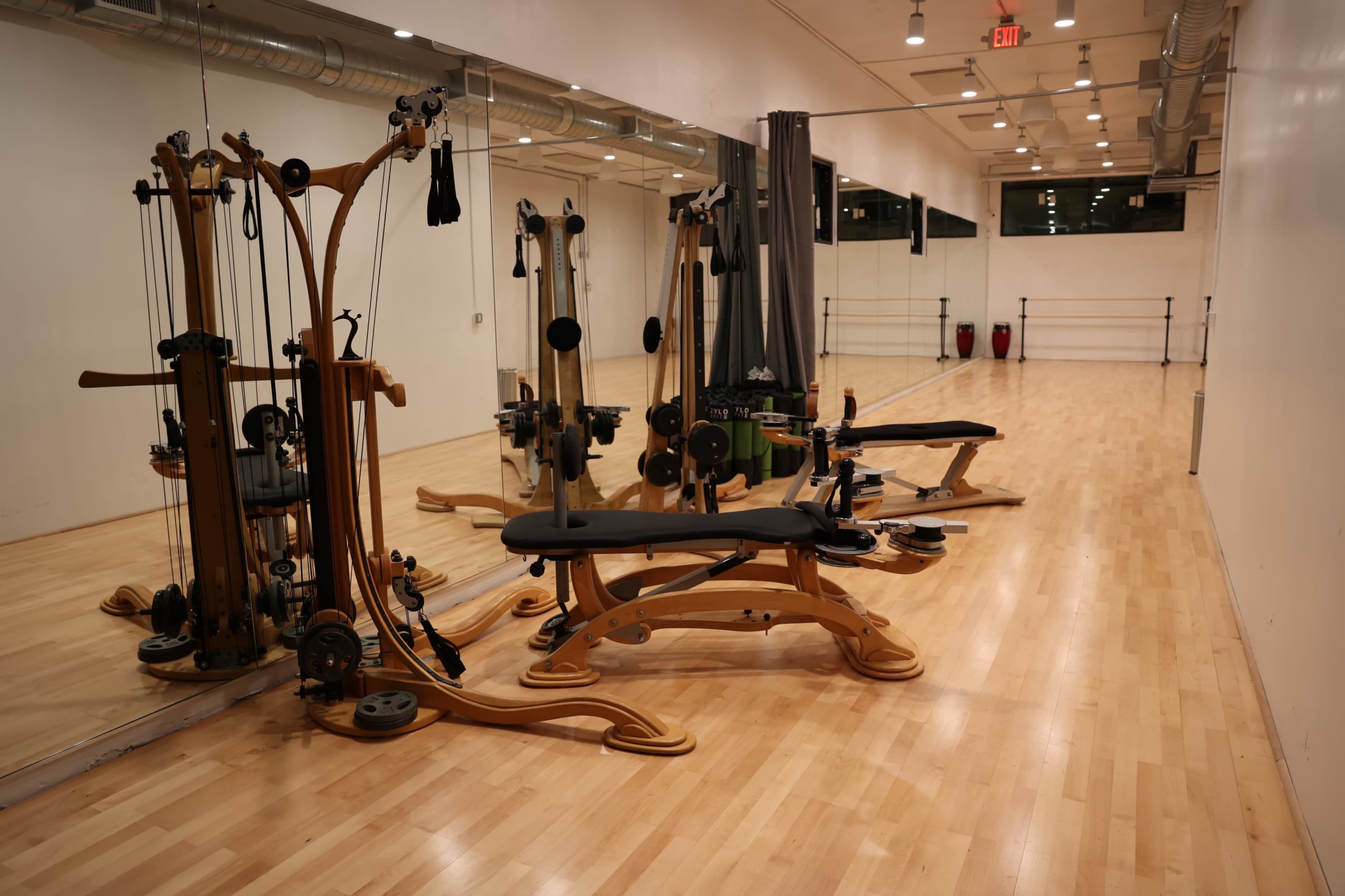 A brightly lit exercise studio with wooden flooring and several gym equipment pieces, including a reformer and cables, along one wall.