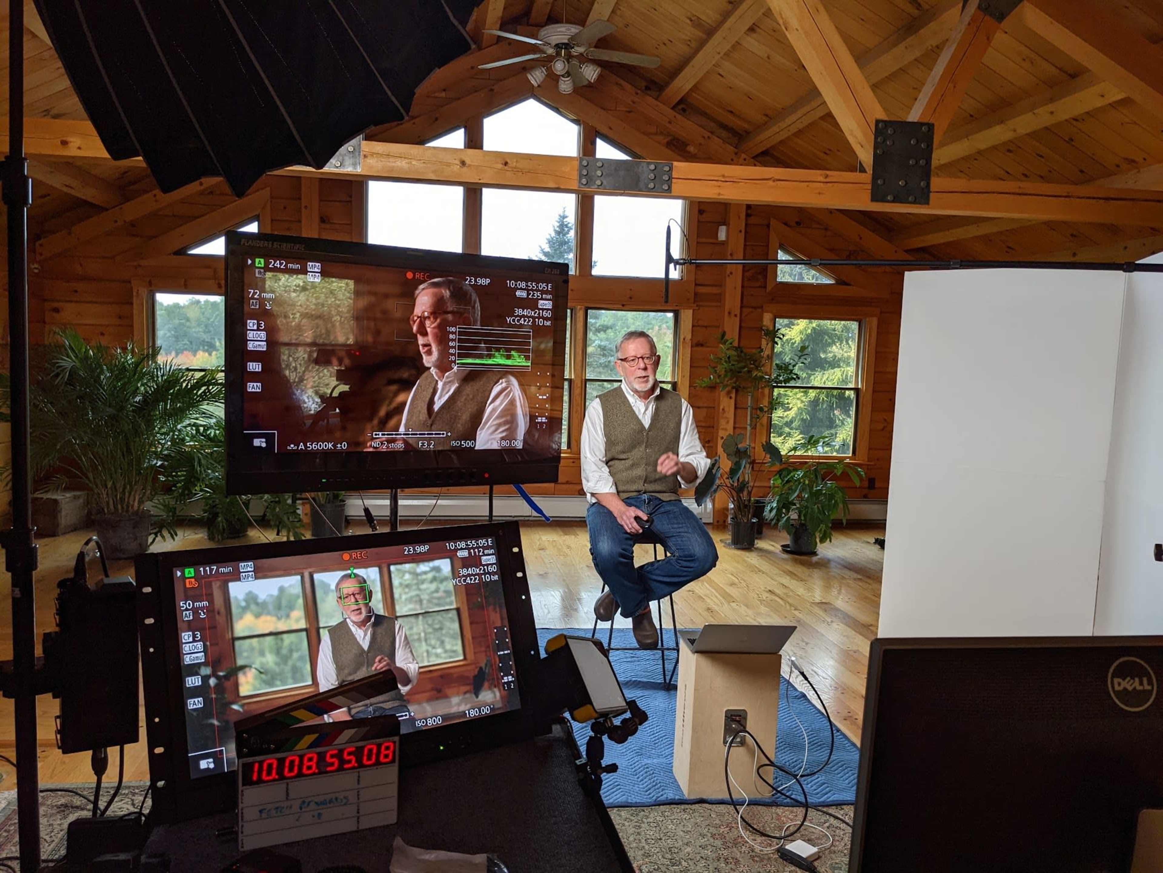 A man wearing a vest sits on a stool in a wooden room with large windows, while a screen behind him displays video footage of the same scene.