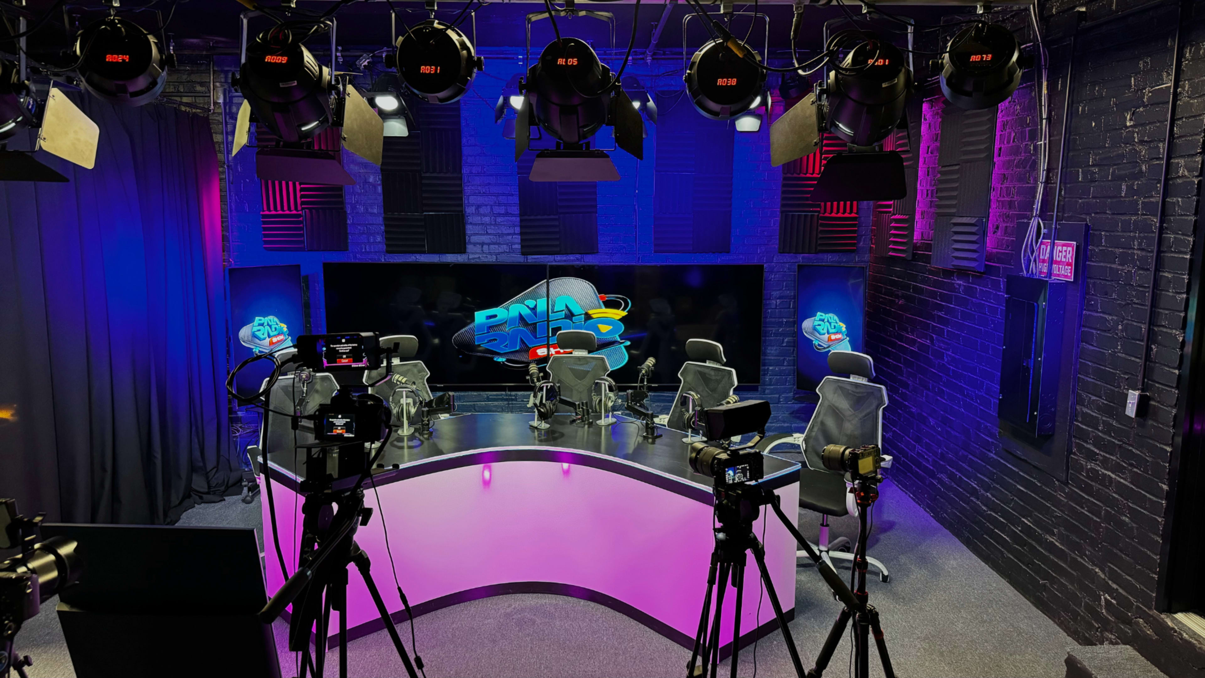 A modern studio setup features multiple cameras, bright lighting, and a central table surrounded by chairs.