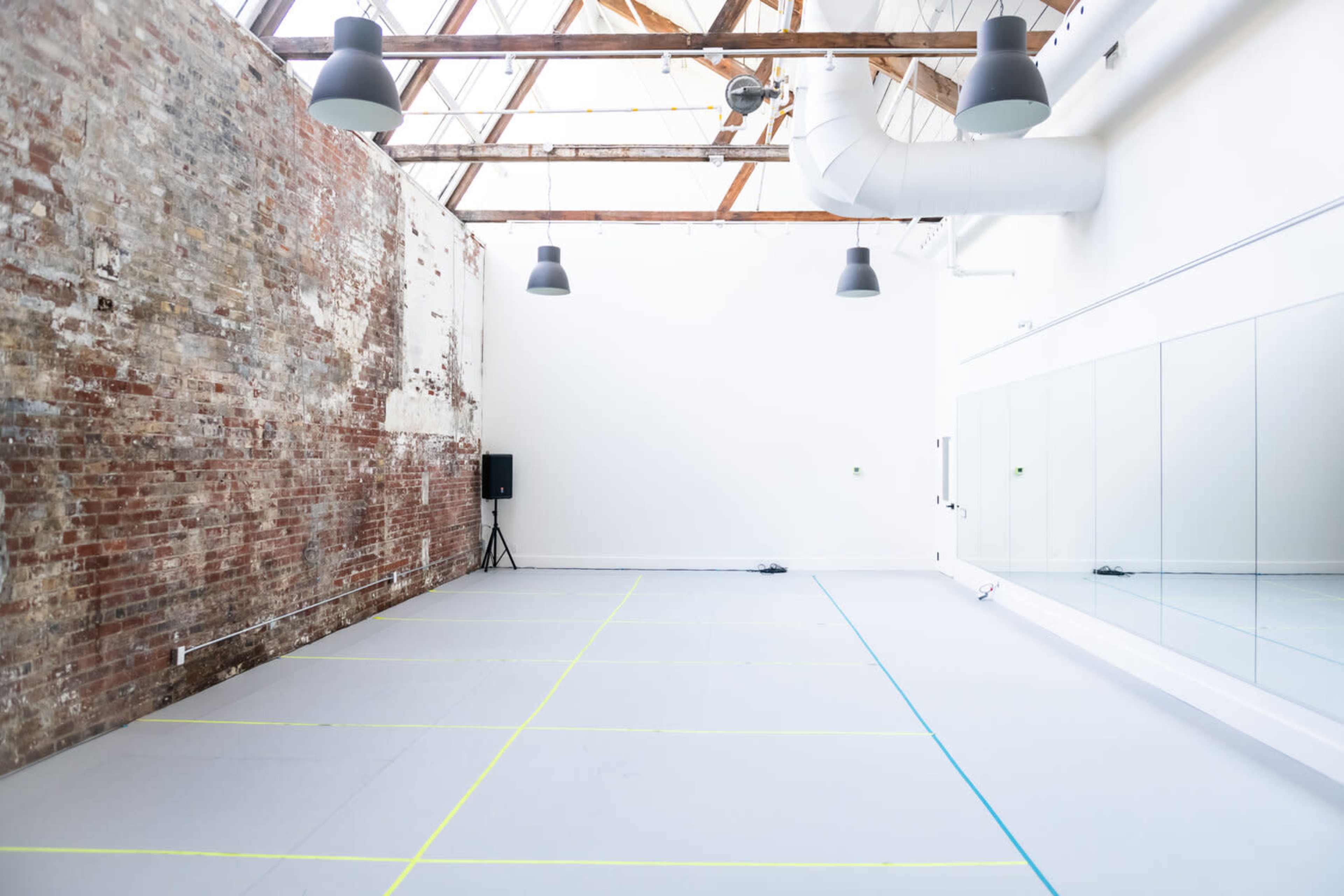 A spacious dance studio with exposed brick walls, a mirrored wall, and a gray floor marked with blue and yellow lines.