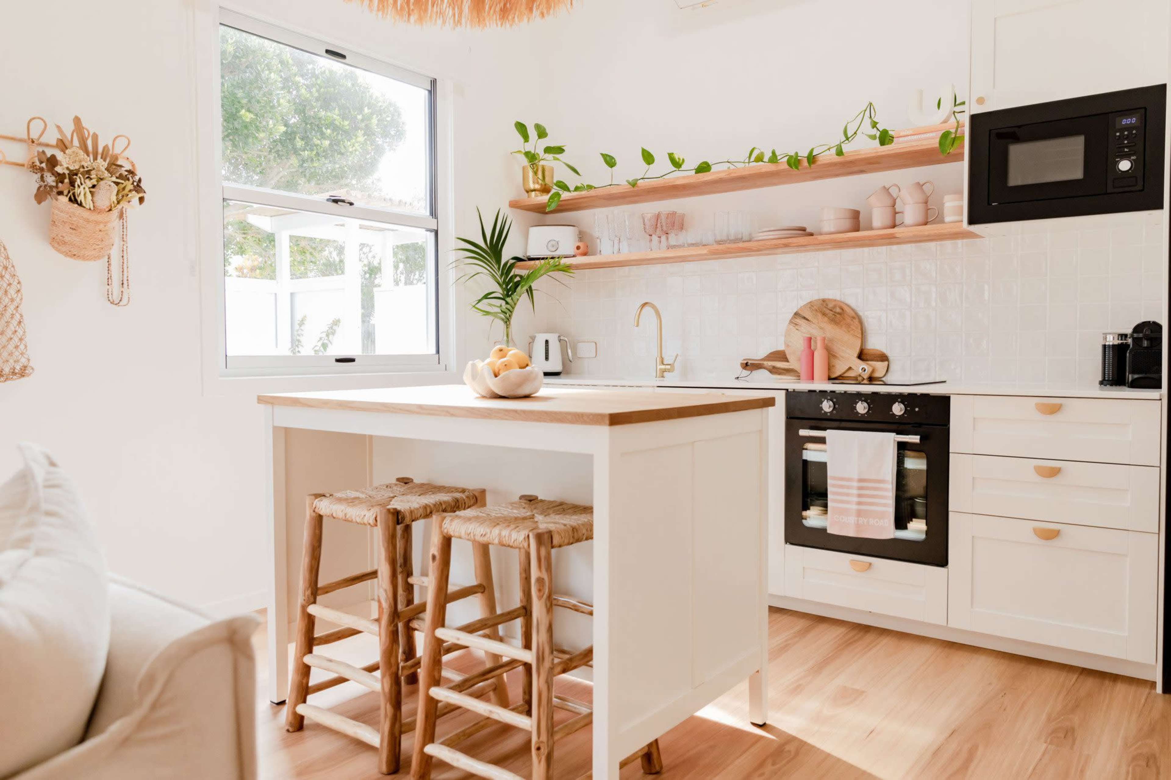 A bright kitchen features open shelving with pink dishware, a wooden island with bar stools, and modern appliances against a white backdrop.