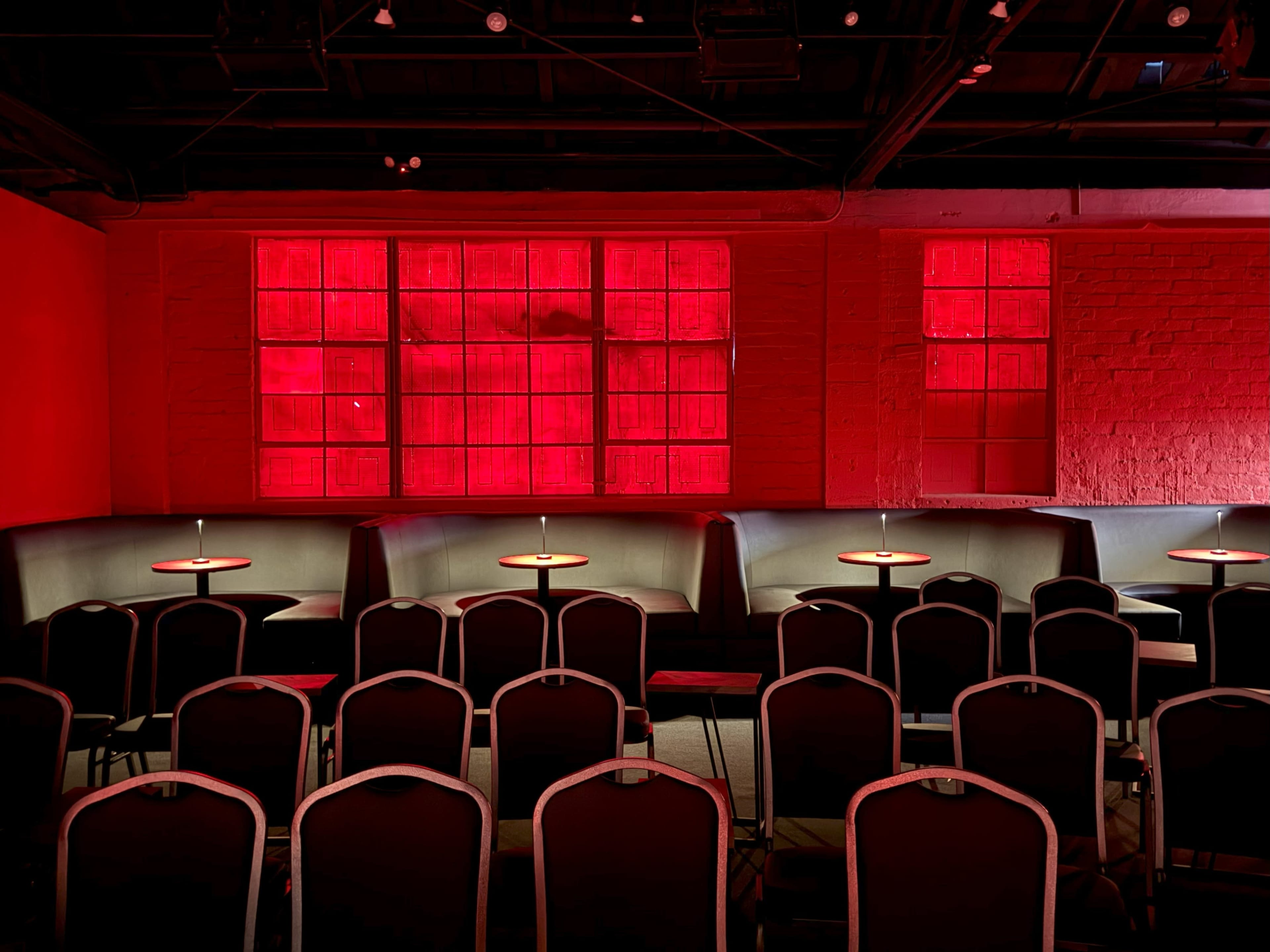 The image shows a dimly lit room with rows of black chairs arranged in front of a series of illuminated red windows.