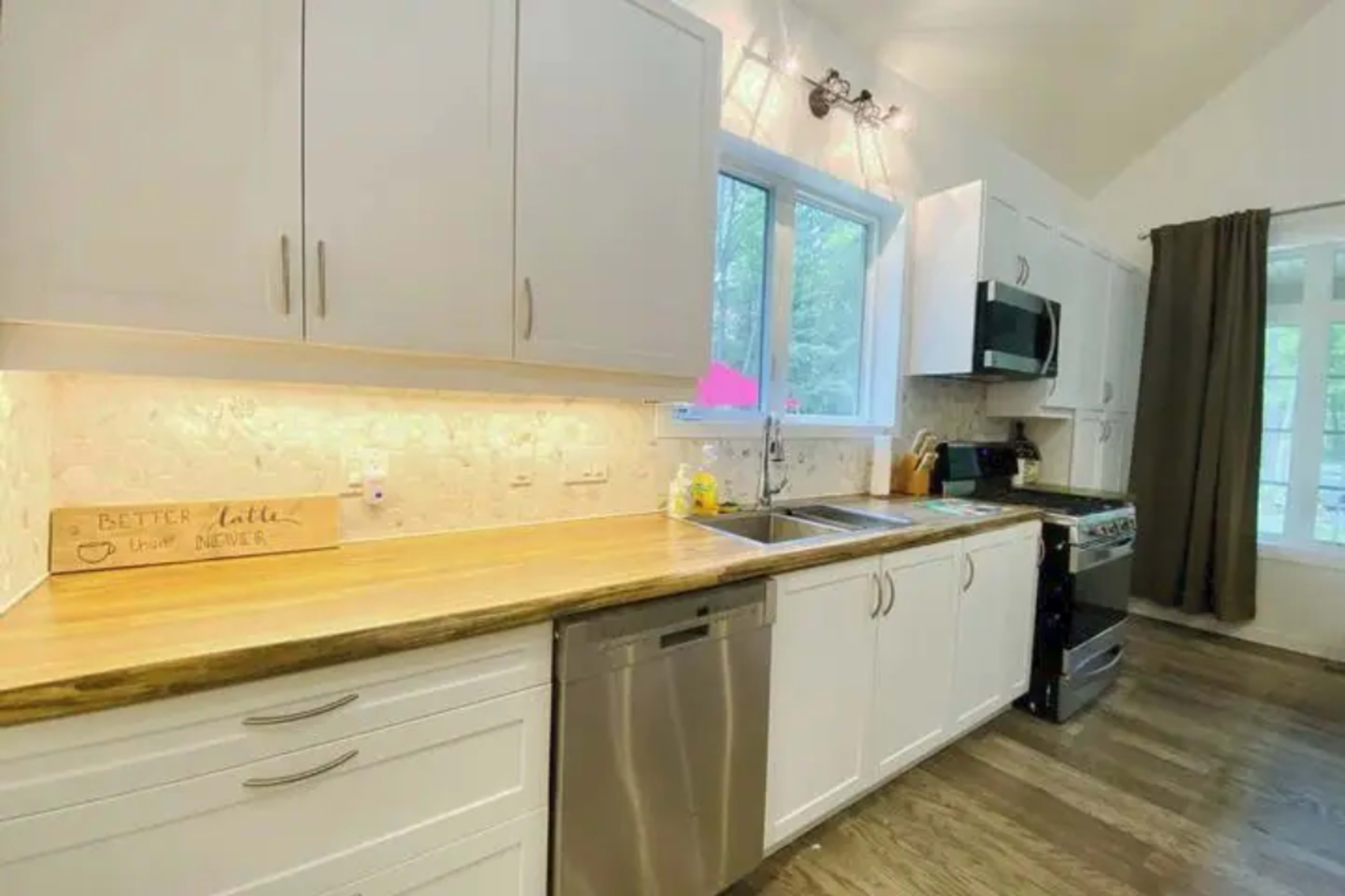 The image shows a modern kitchen featuring white cabinets, a wooden countertop, stainless steel appliances, and a window over the sink.