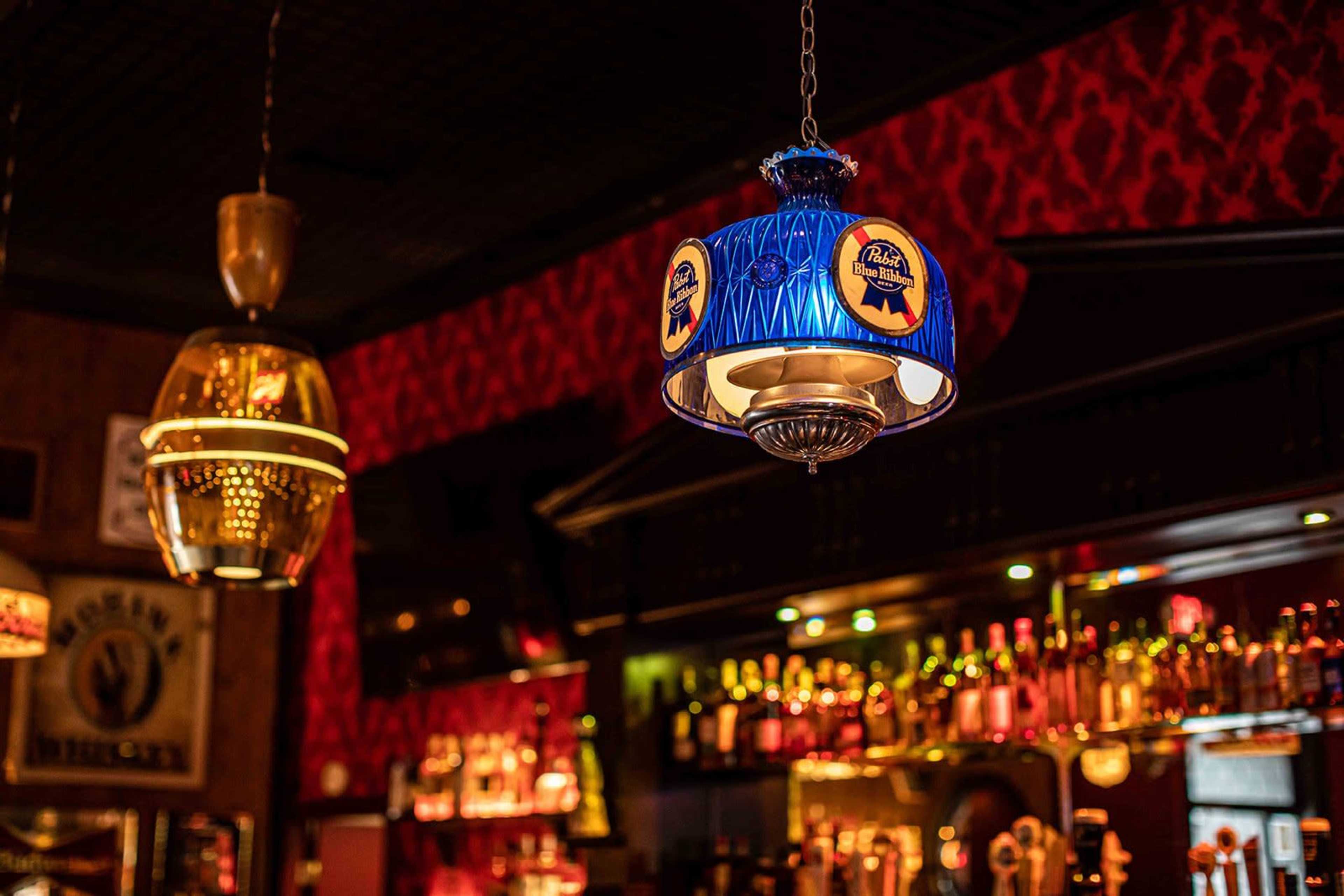 The image shows two decorative pendant lamps hanging from the ceiling in a bar, with a backdrop of illuminated shelves filled with bottles.