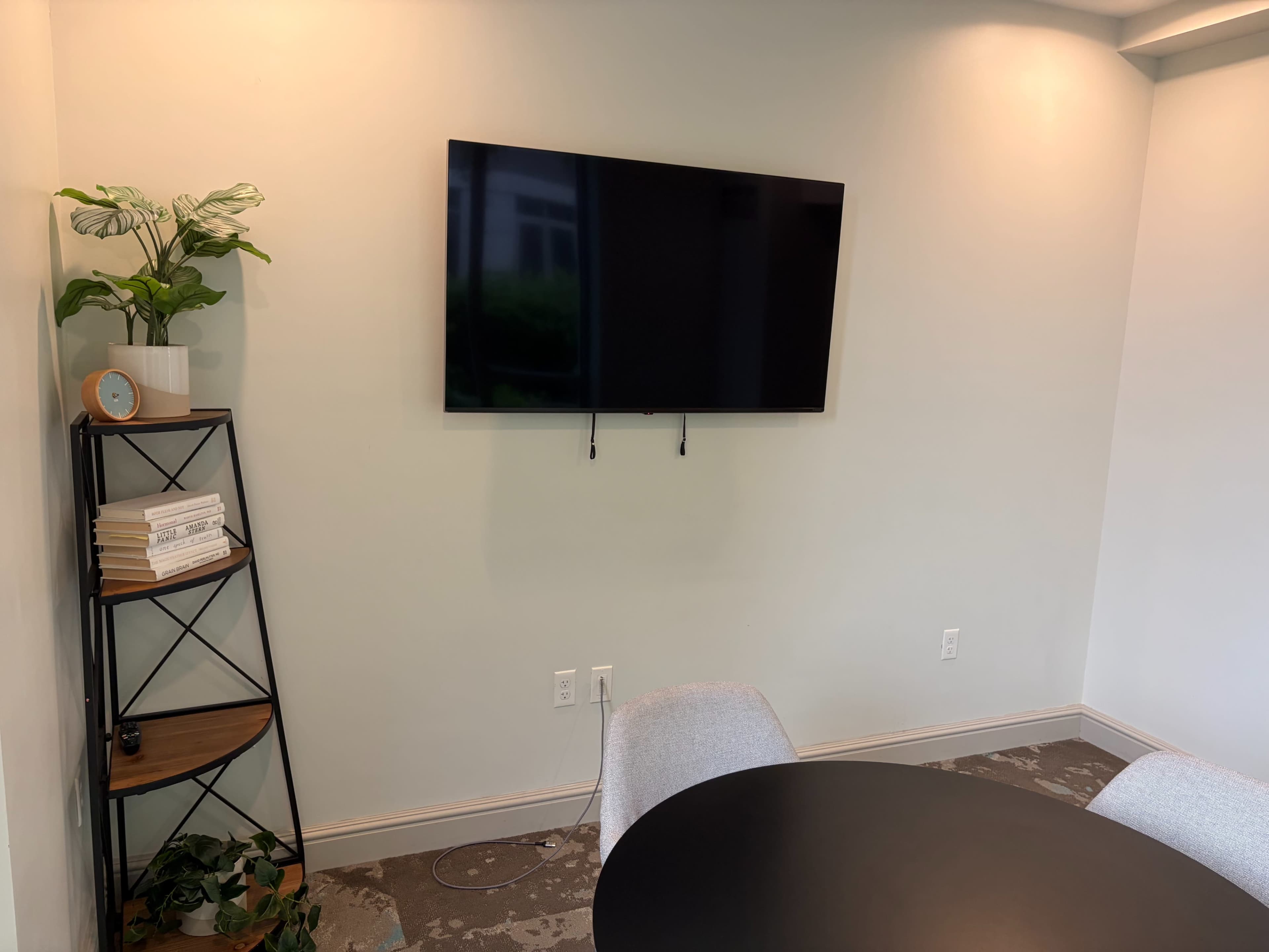 The image shows a small meeting area with a wall-mounted TV, a black round table, gray chairs, and a plant on a shelf.