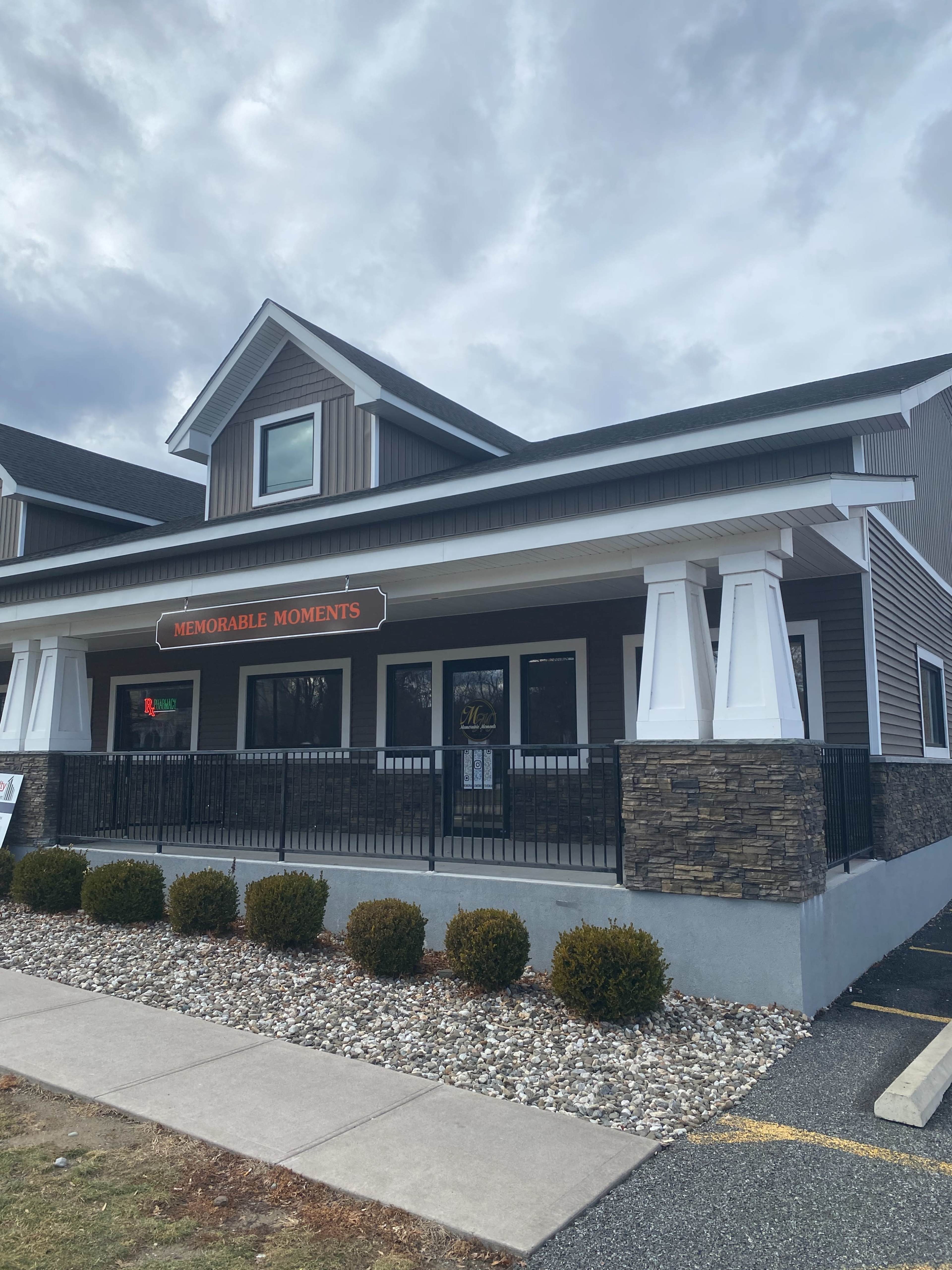 The image shows a two-story building with a porch, featuring a sign that reads "Memorable Moments" above the entrance, surrounded by landscaped bushes and a gravel area.