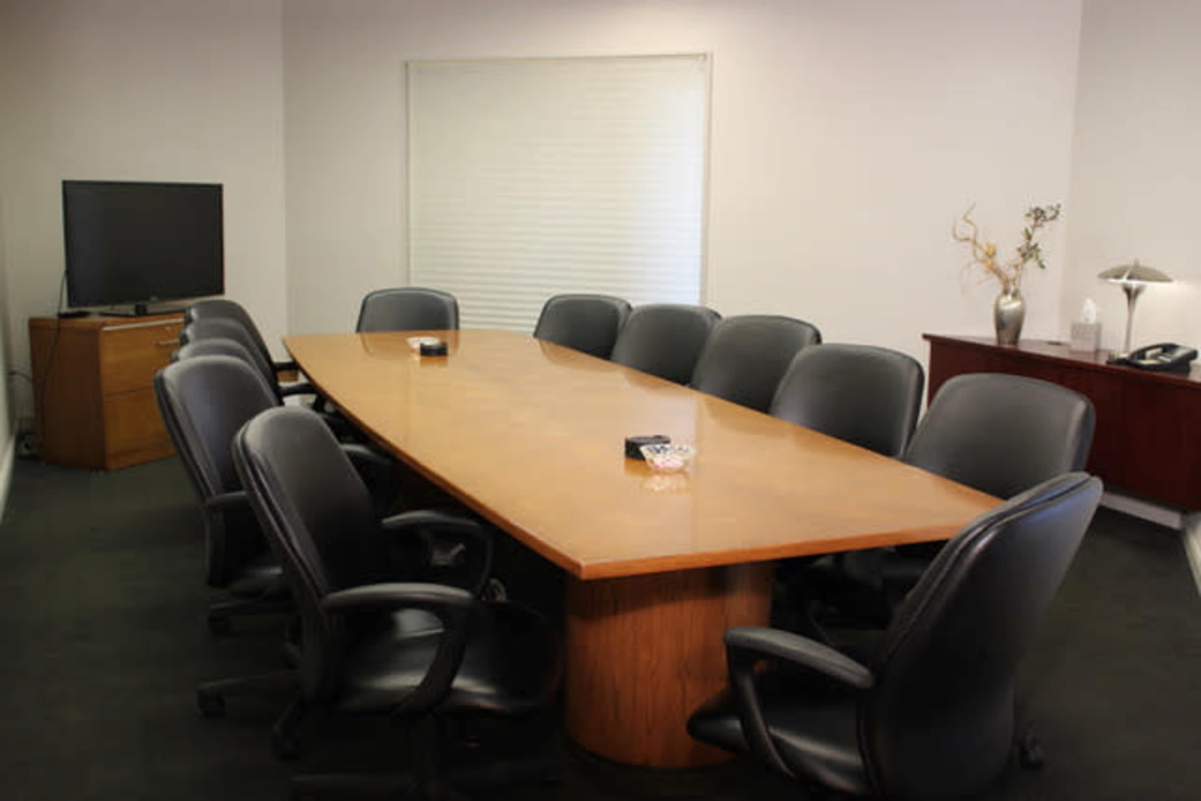 A conference room features a long wooden table surrounded by black ergonomic chairs, with a window and television in the background.