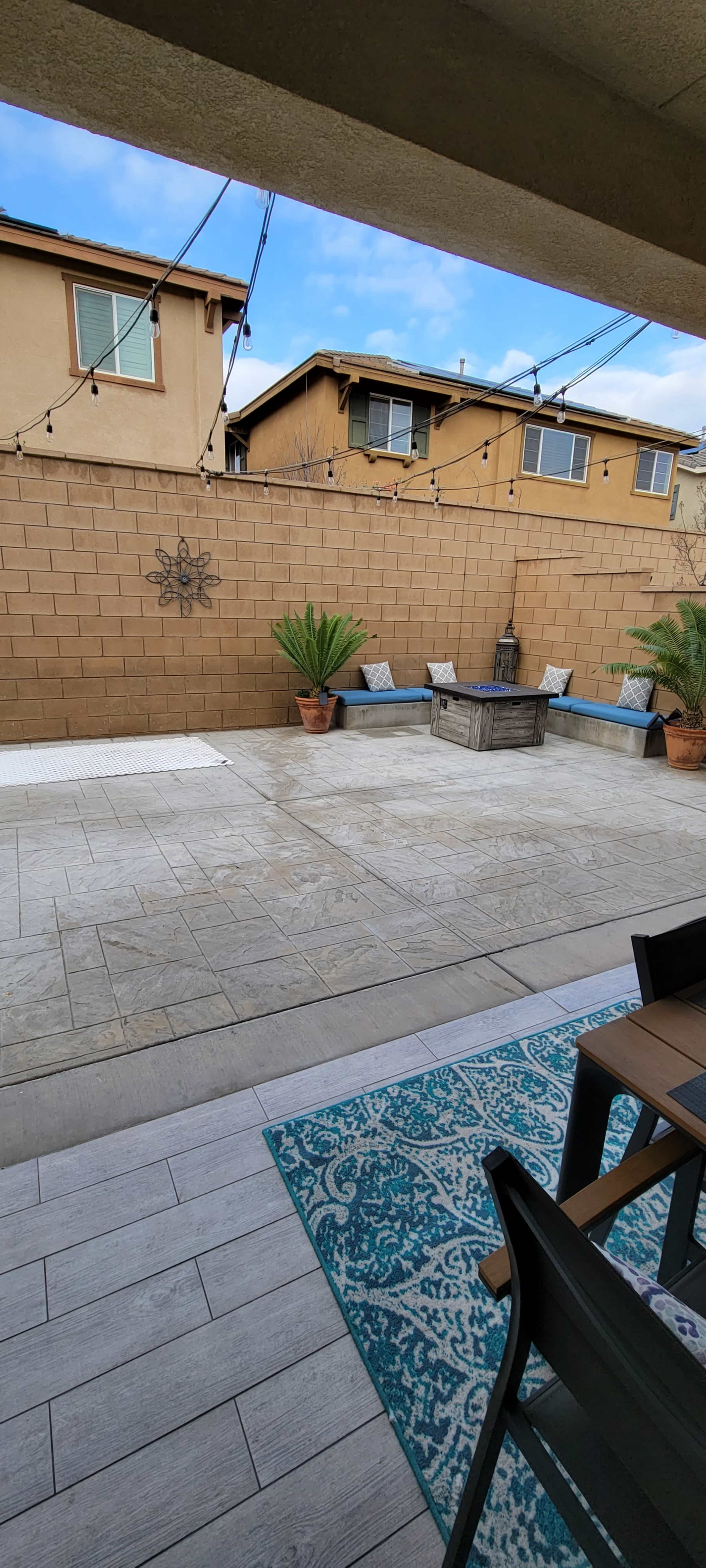The image shows a landscaped patio area with a concrete floor, potted plants, and a decorative wall, with string lights overhead and a seating arrangement visible.