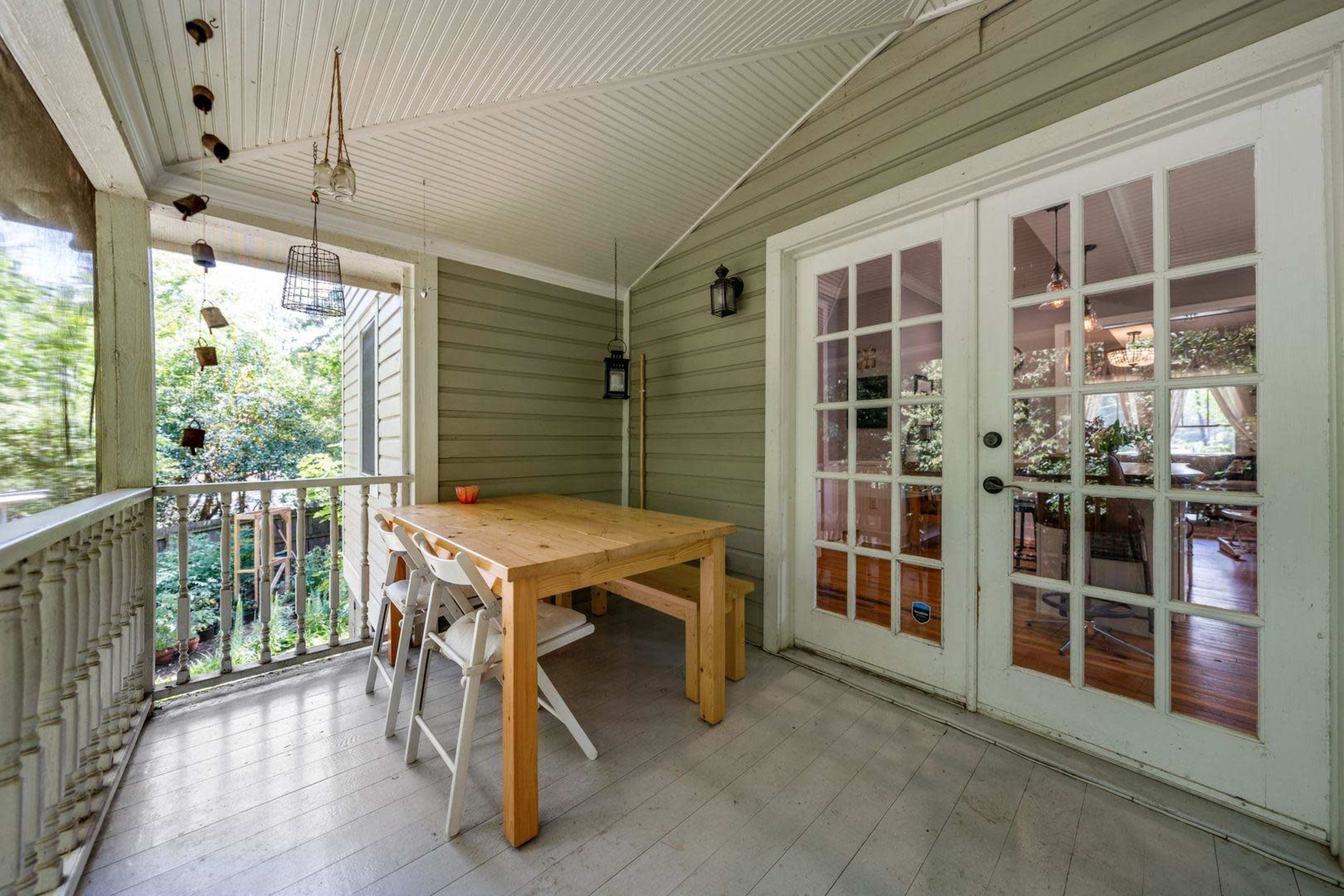 A wooden table with white chairs is positioned on a covered porch next to a set of French doors leading to an interior space with visible greenery outside.