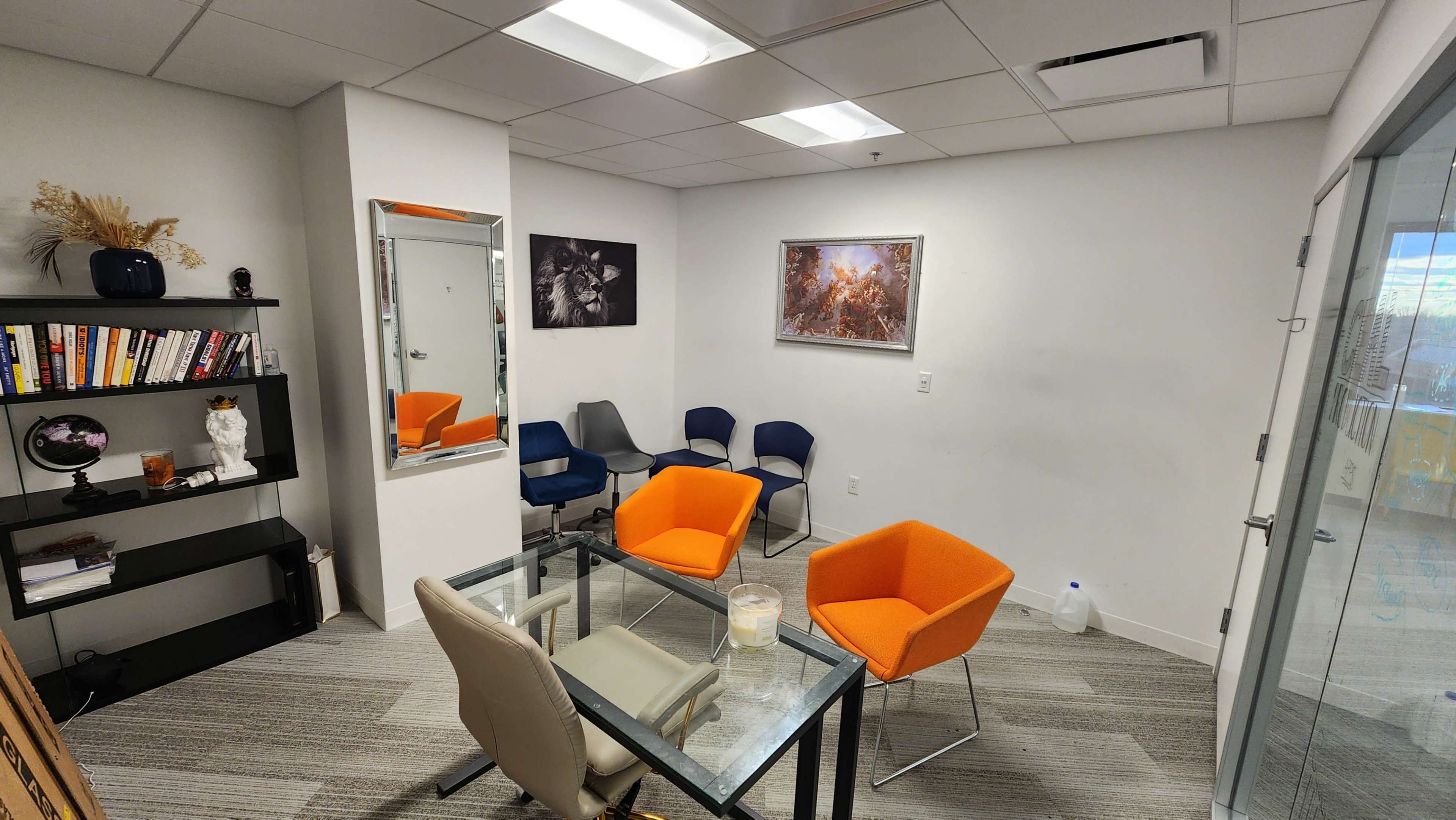 A small office meeting room features a glass table with orange and blue chairs, a bookshelf filled with books, and wall decorations including a mirror and framed artwork.