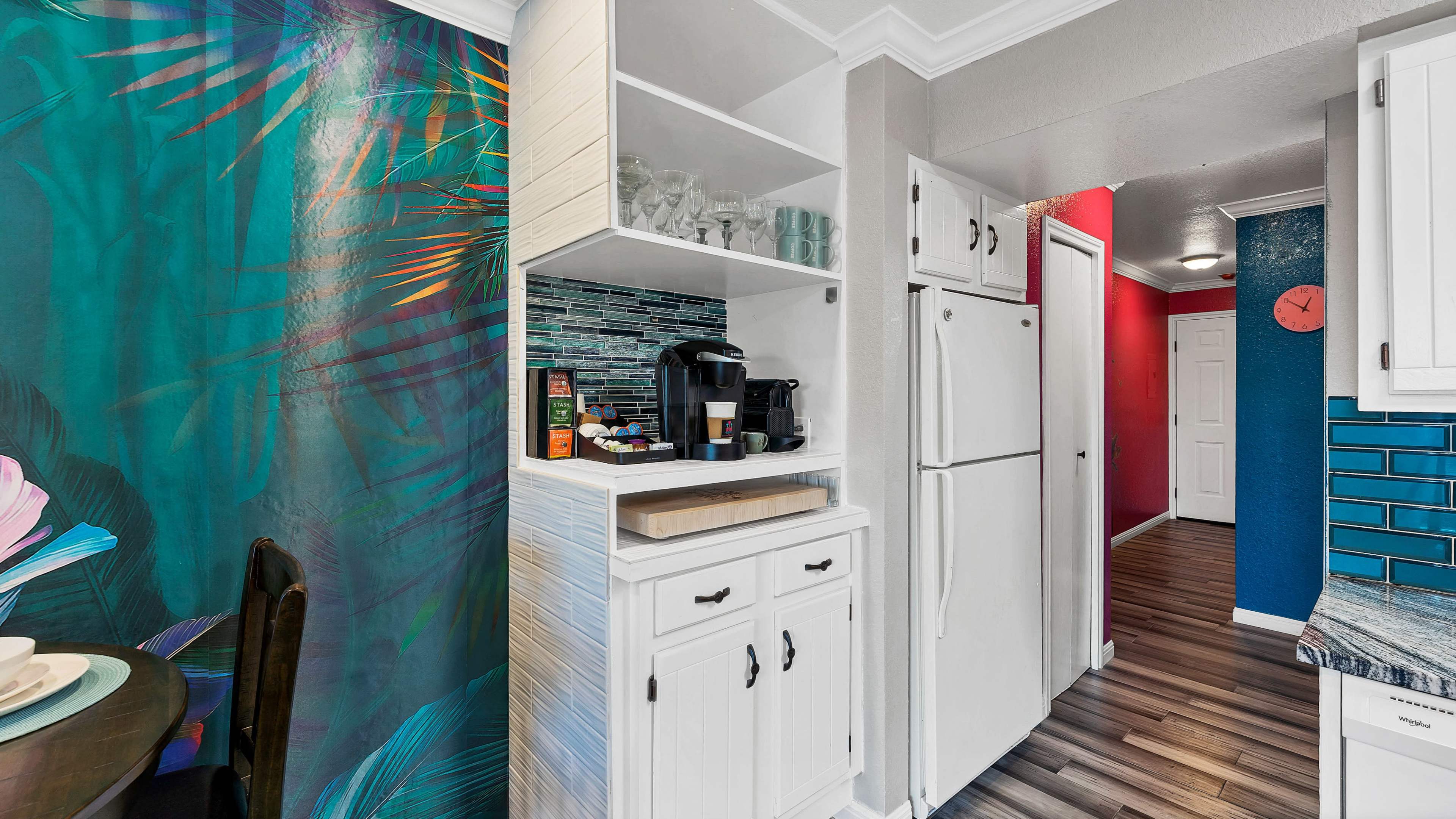 A brightly colored kitchen with a tropical-themed wall mural, open shelving displaying glassware and a coffee maker, and a hallway leading to a white door.