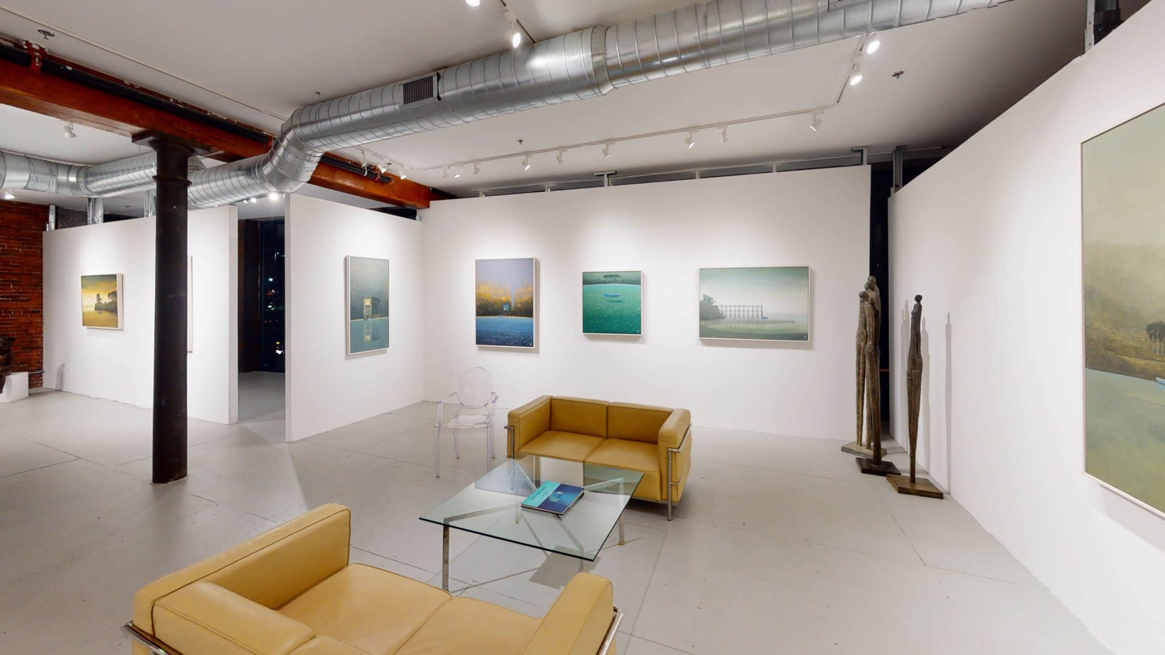A modern art gallery with white walls displaying framed paintings, featuring a seating area with a glass table and yellow couches.