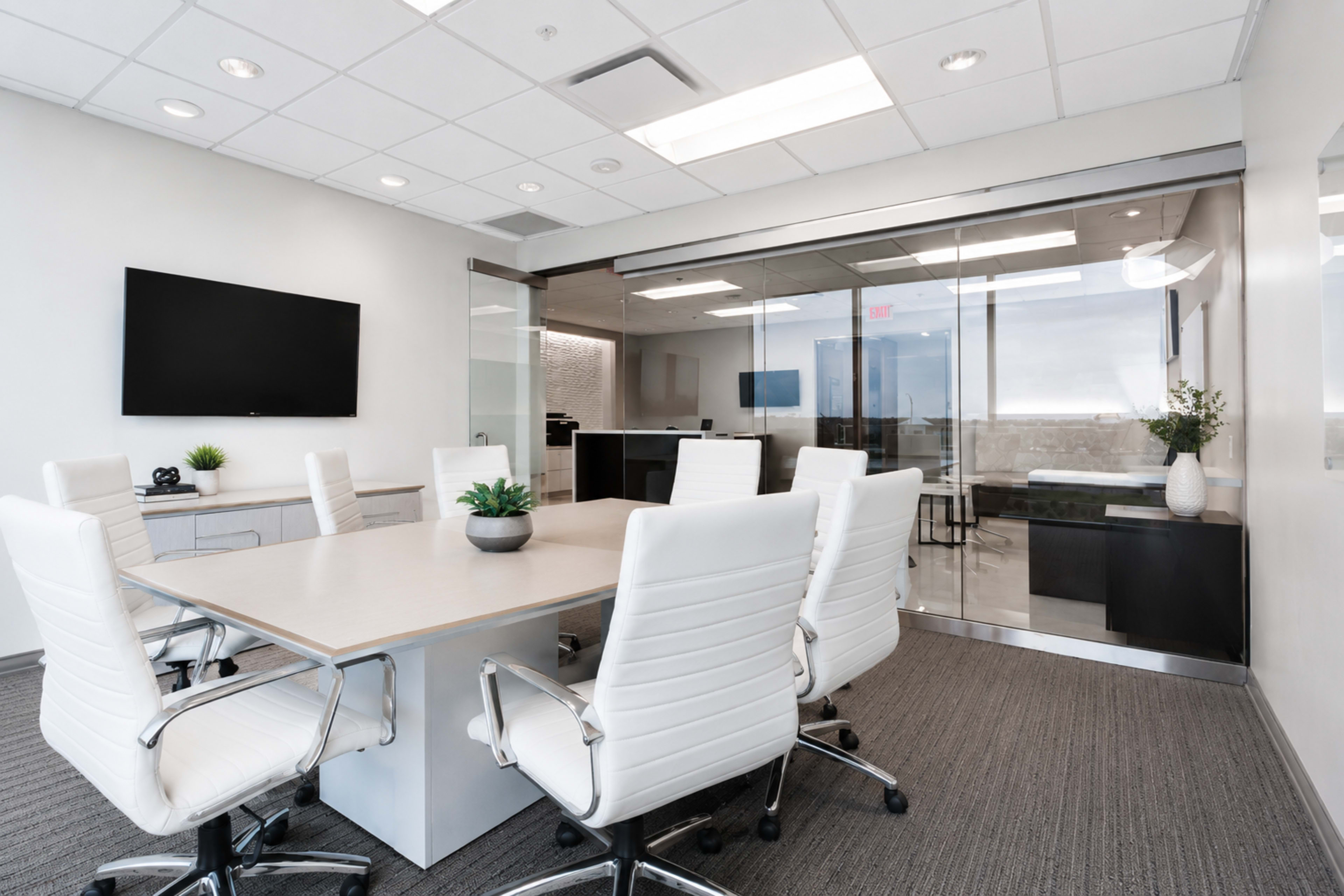 The image shows a modern conference room with a large table surrounded by white ergonomic chairs and a glass-walled area that connects to another space.