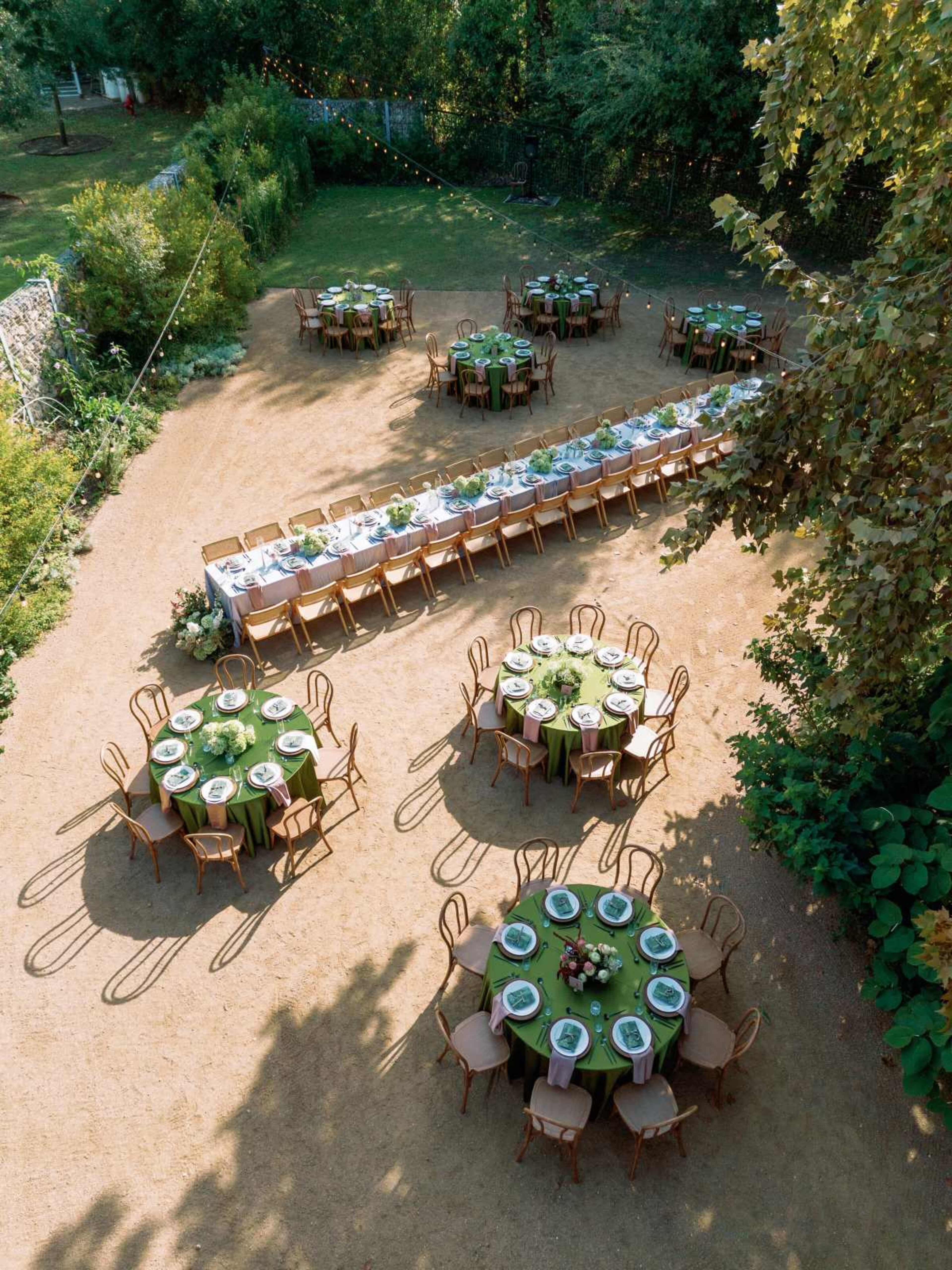 The scene shows an outdoor dining area with multiple tables set for a gathering, surrounded by greenery and a dirt pathway.