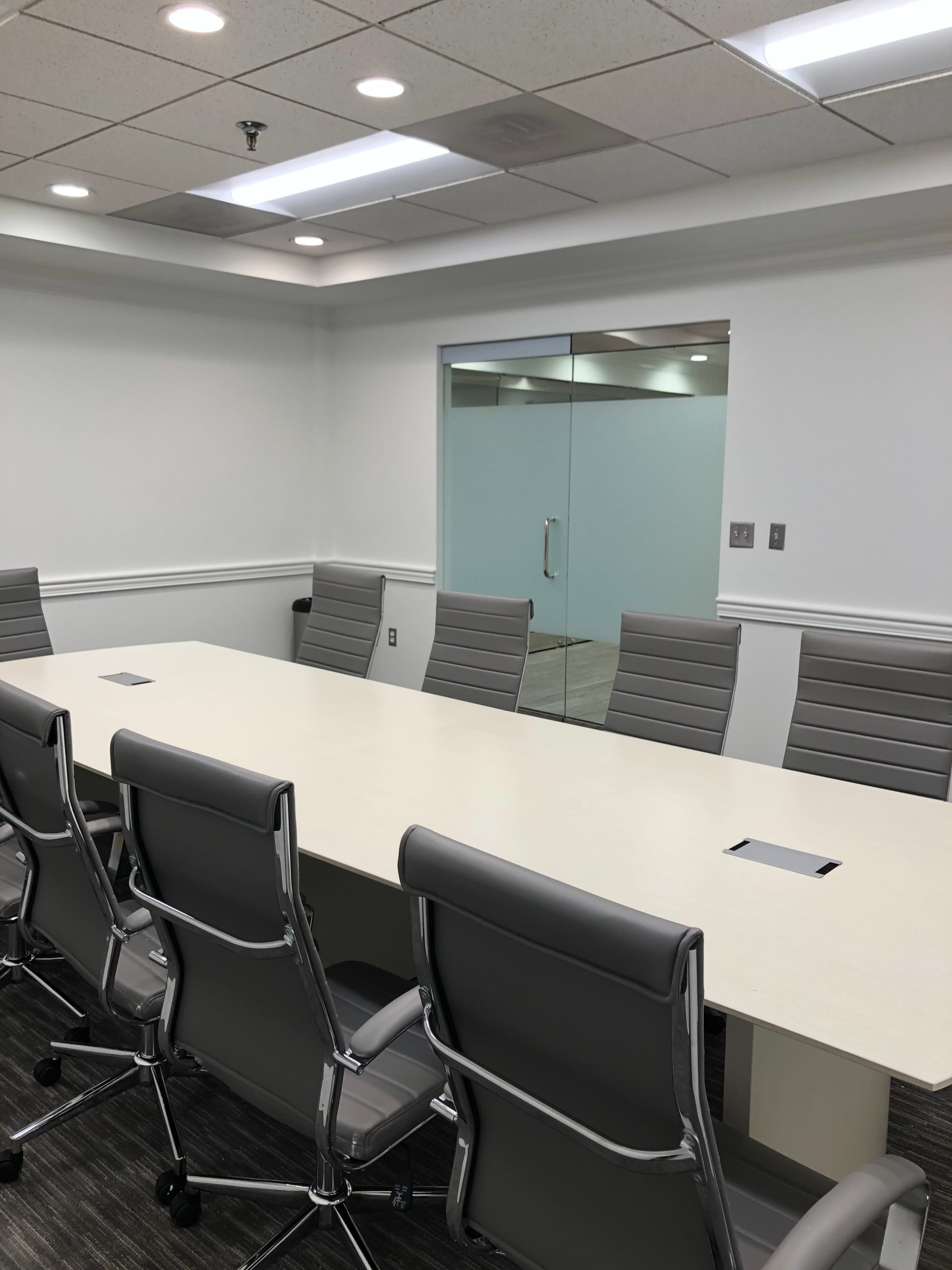 A modern conference room with a long white table and gray ergonomic chairs, featuring a glass door leading to another room.