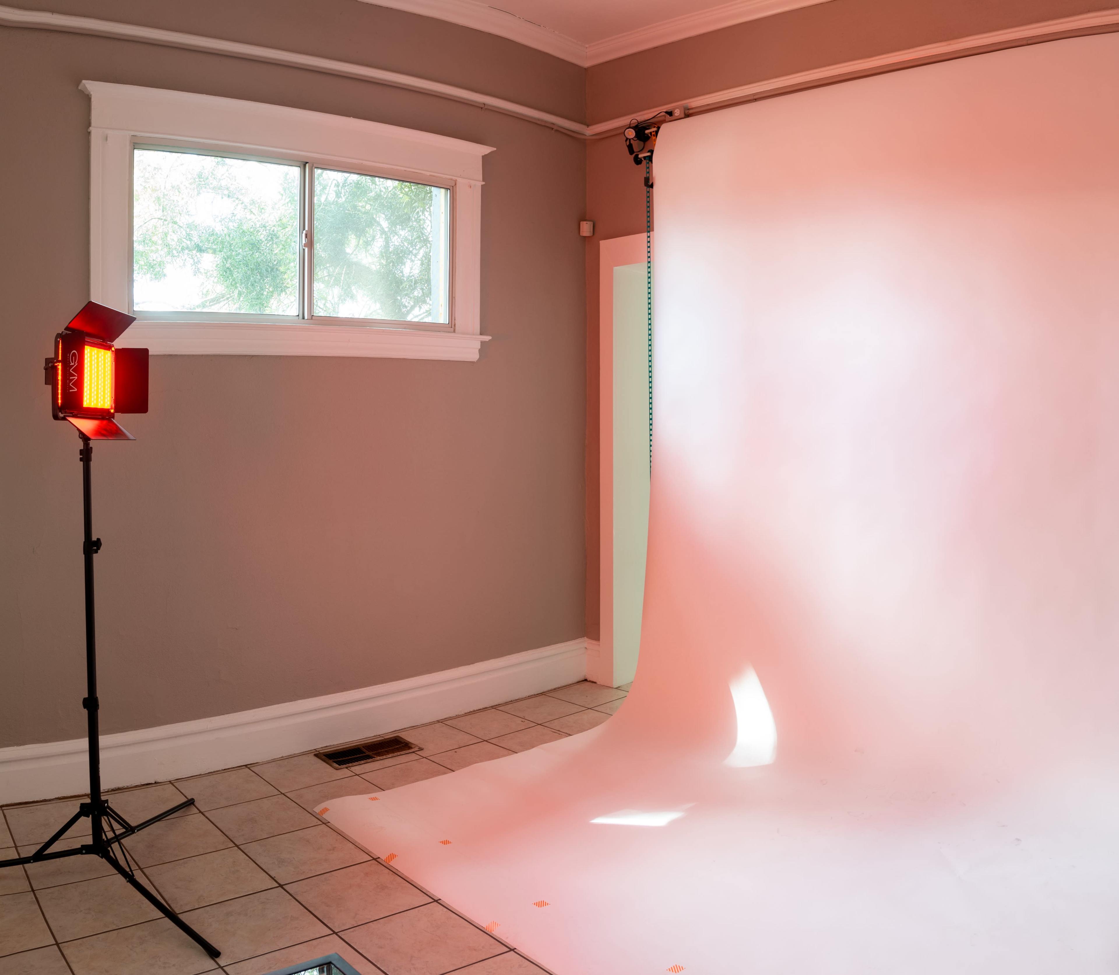 A photography studio setup with a roll of white backdrop, a window, and a lighting device positioned nearby.
