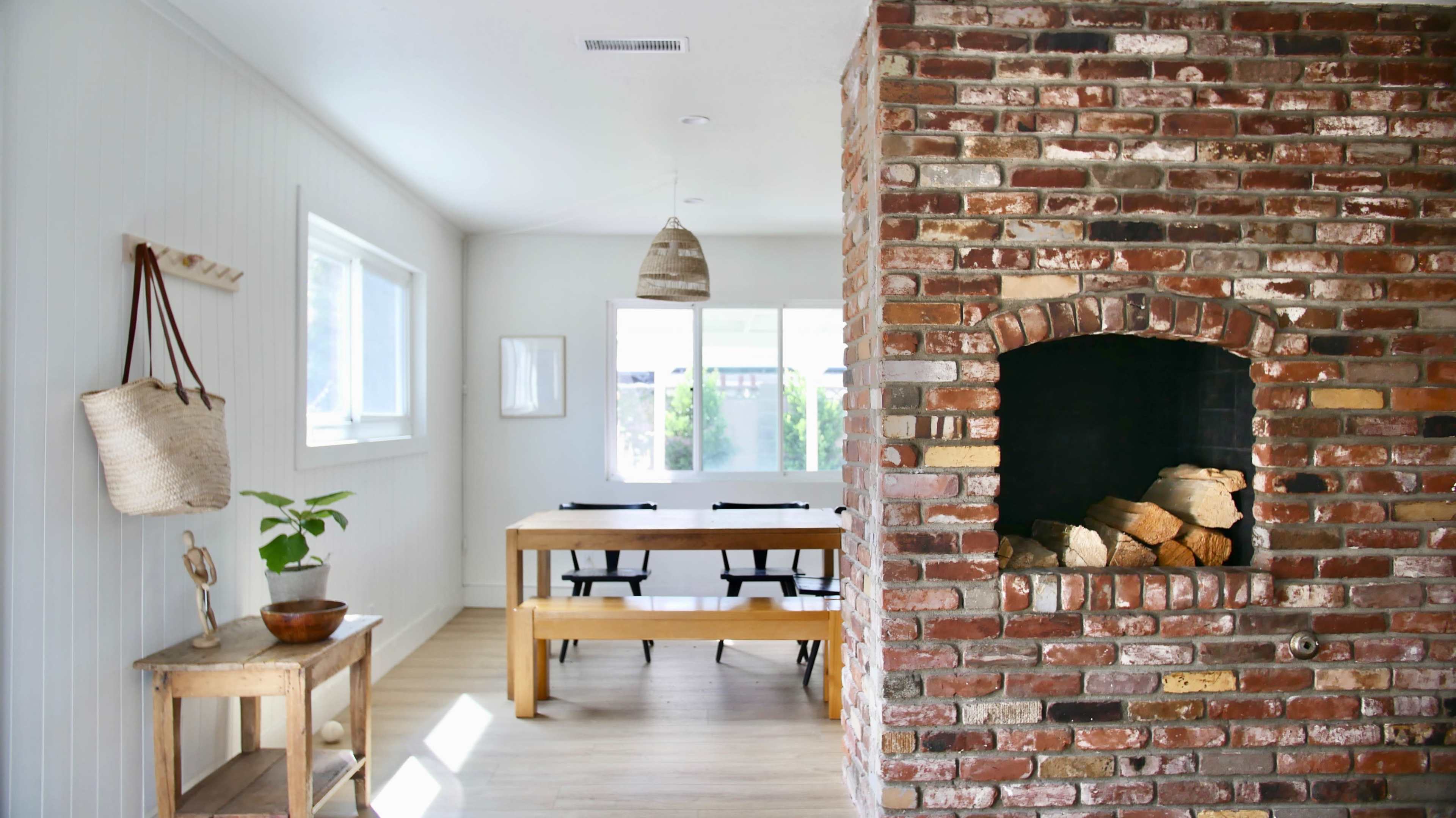 A spacious room features a brick fireplace next to a wooden dining table surrounded by chairs and large windows.