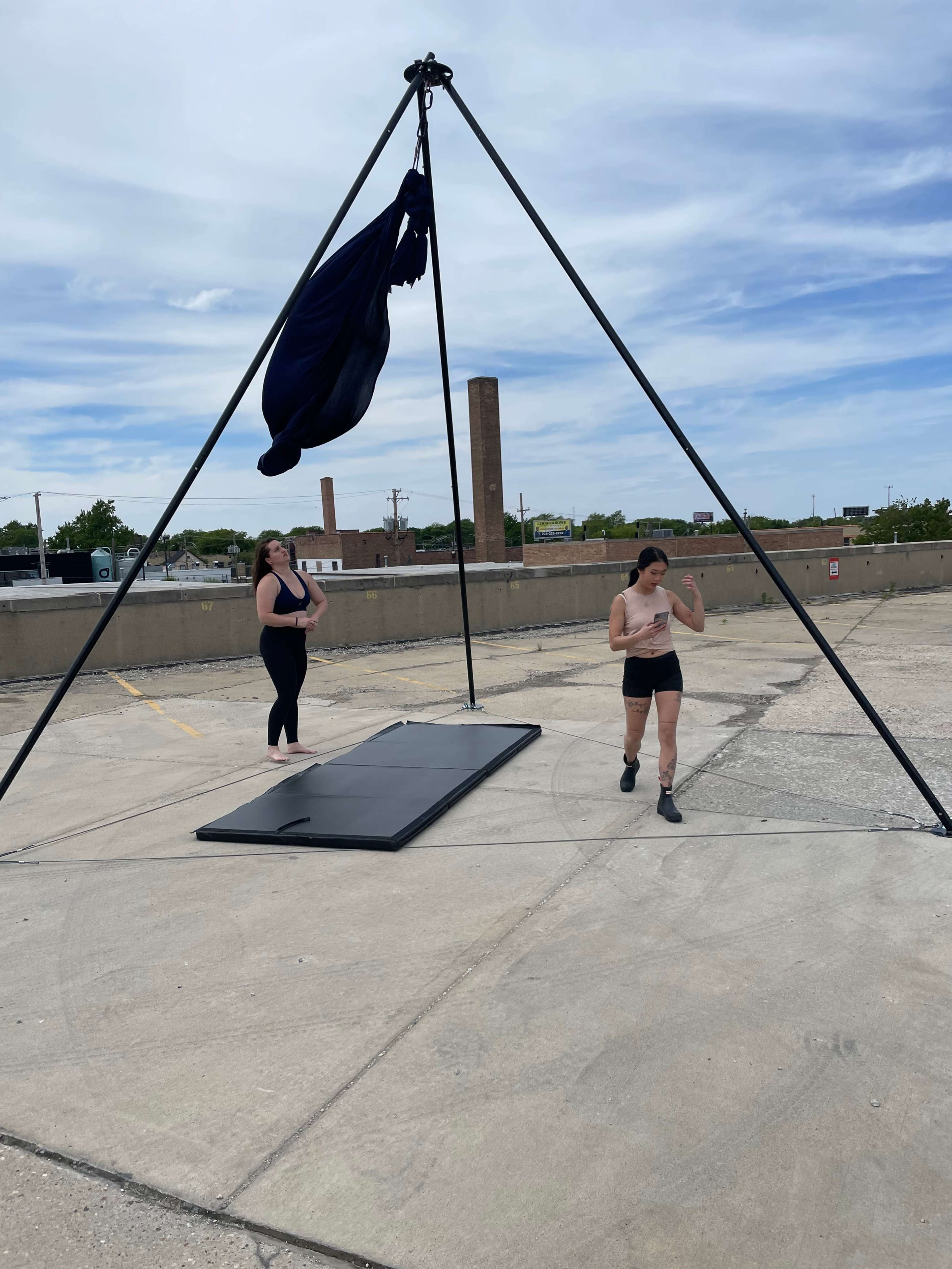 Two individuals engage in aerial arts practice using a black fabric suspended from a tripod frame on a rooftop.